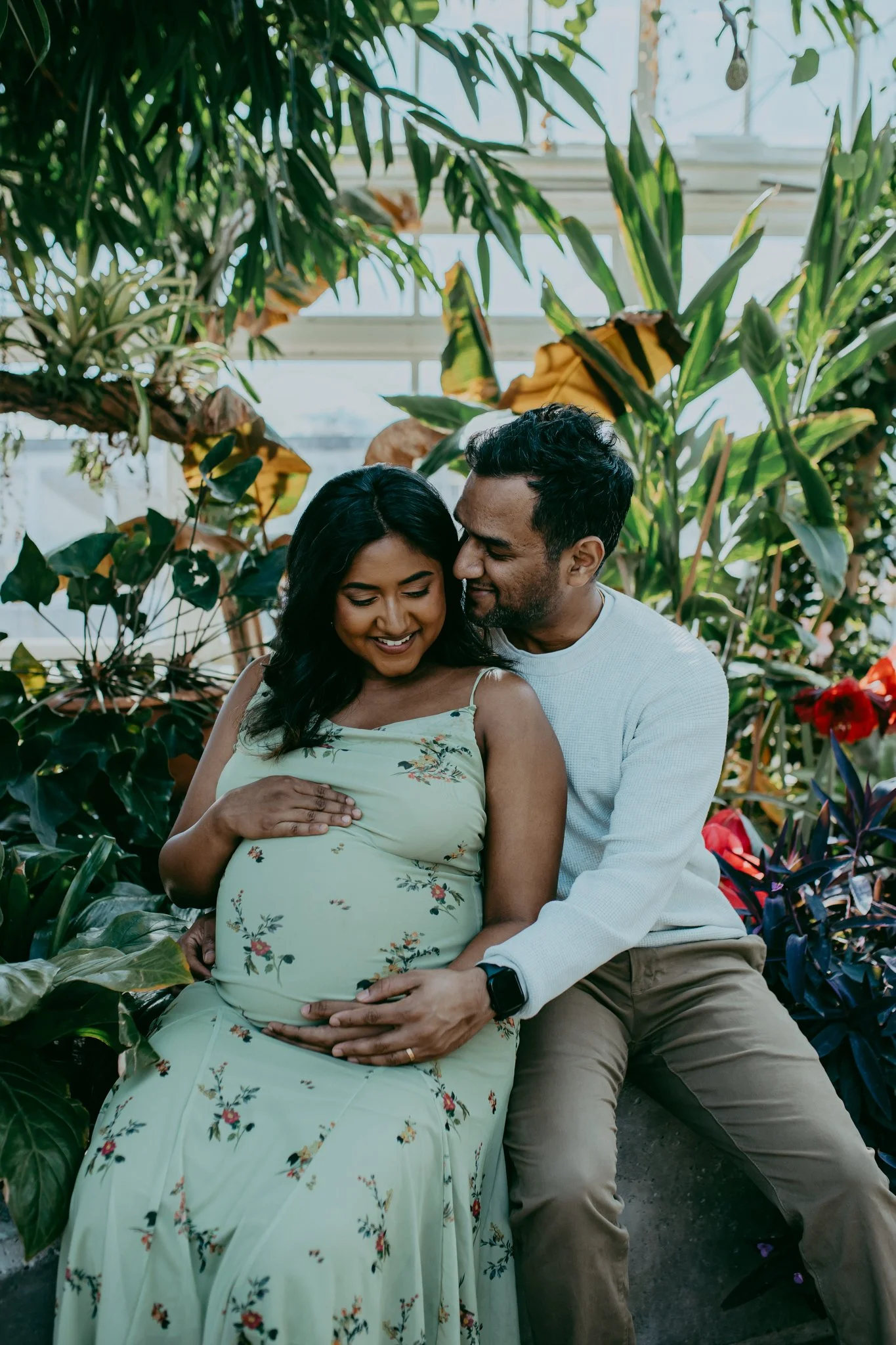A pregnant woman and a man sit close together in a lush indoor garden, smiling and touching her belly with her hands.