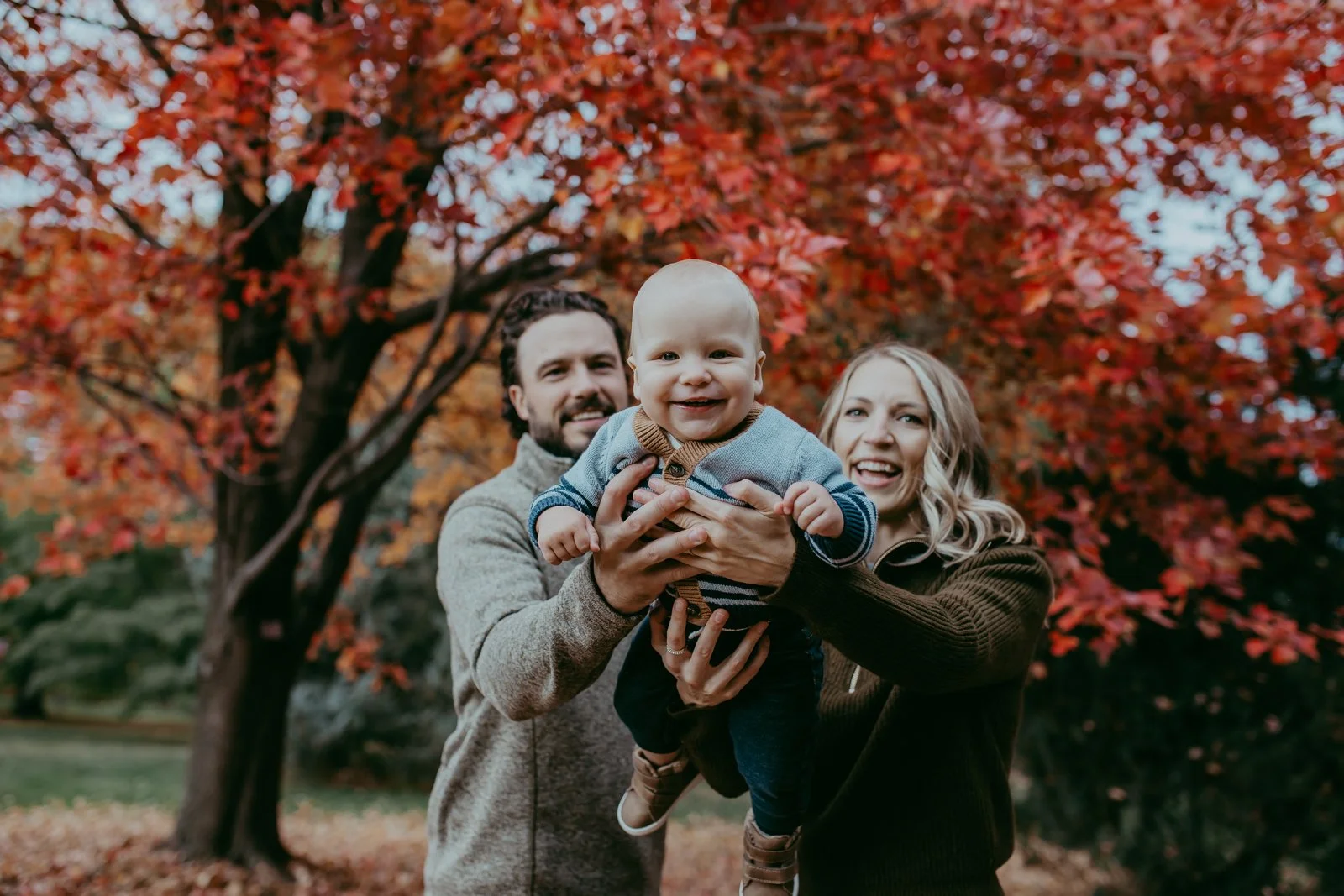 A happy family of three, a man, woman, and baby, enjoying the outdoors during fall with red and orange leaves on trees in the background.