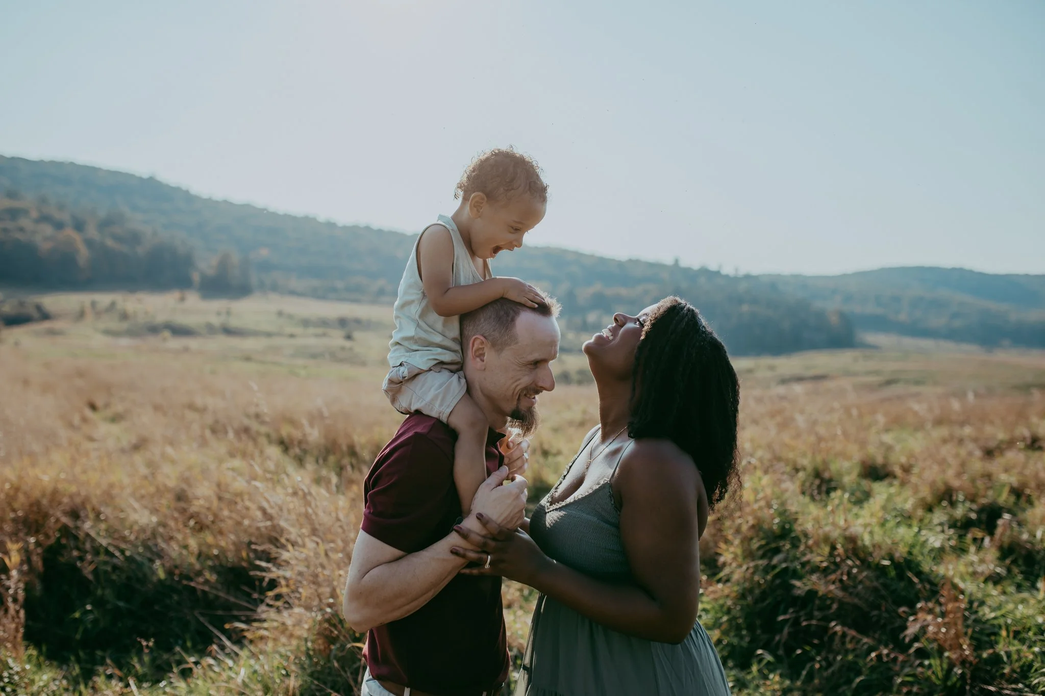 Boy on dad's shoulders, mom looking at son. In a vast field with hills in the background