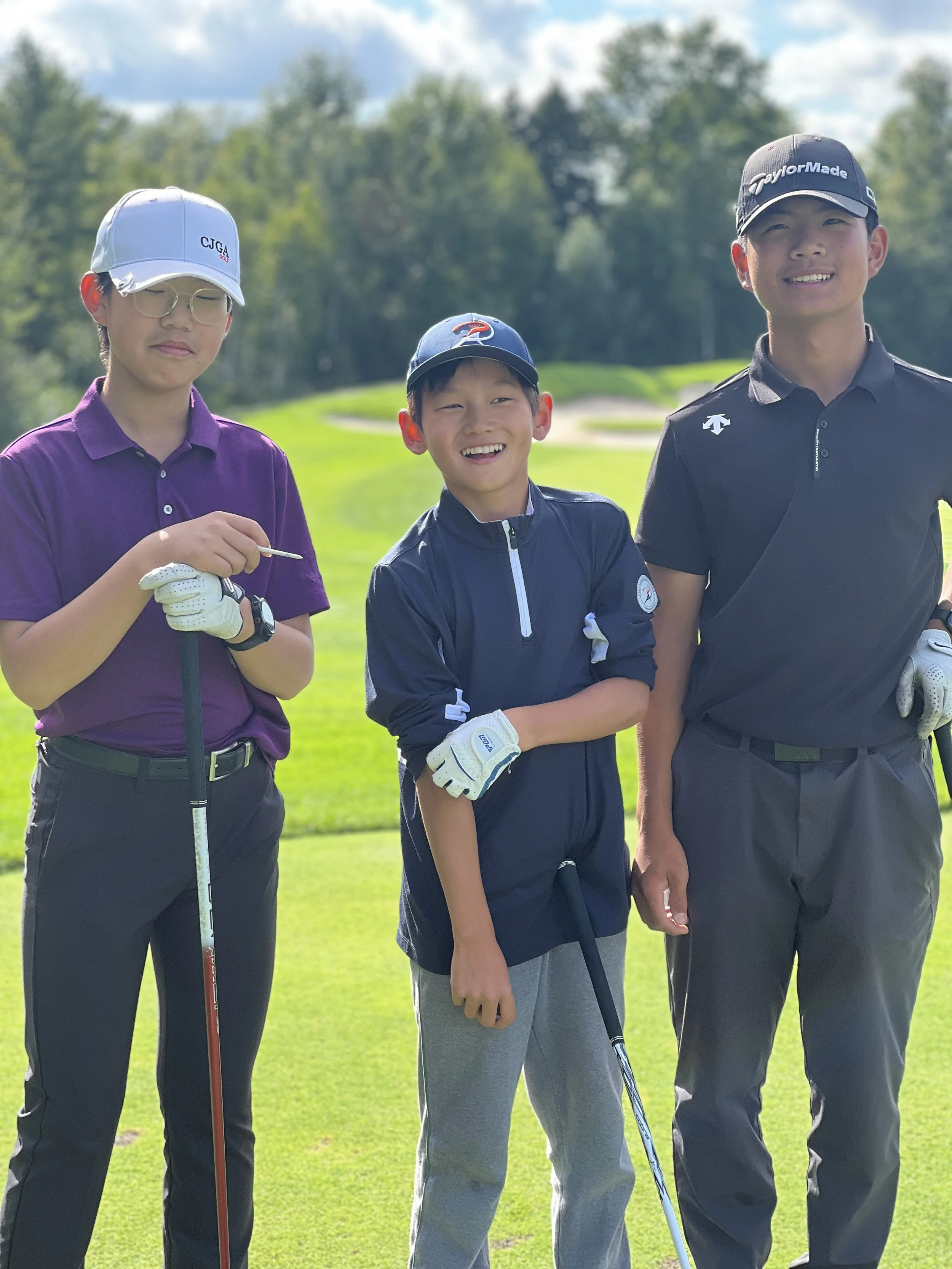 Three boys standing on a golf course, smiling, holding golf clubs, with trees and a blue sky in the background.