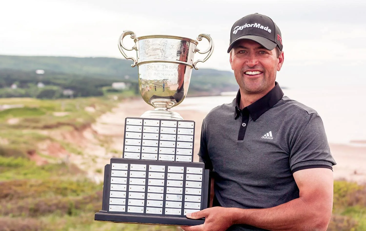 A man wearing a black TaylorMade cap and gray Adidas polo shirt smiling while holding a large trophy and a board with multiple small white plaques outdoors near a coastline.