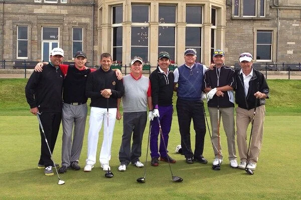 Group of eight men standing on a golf course holding golf clubs, with a historic building in the background.