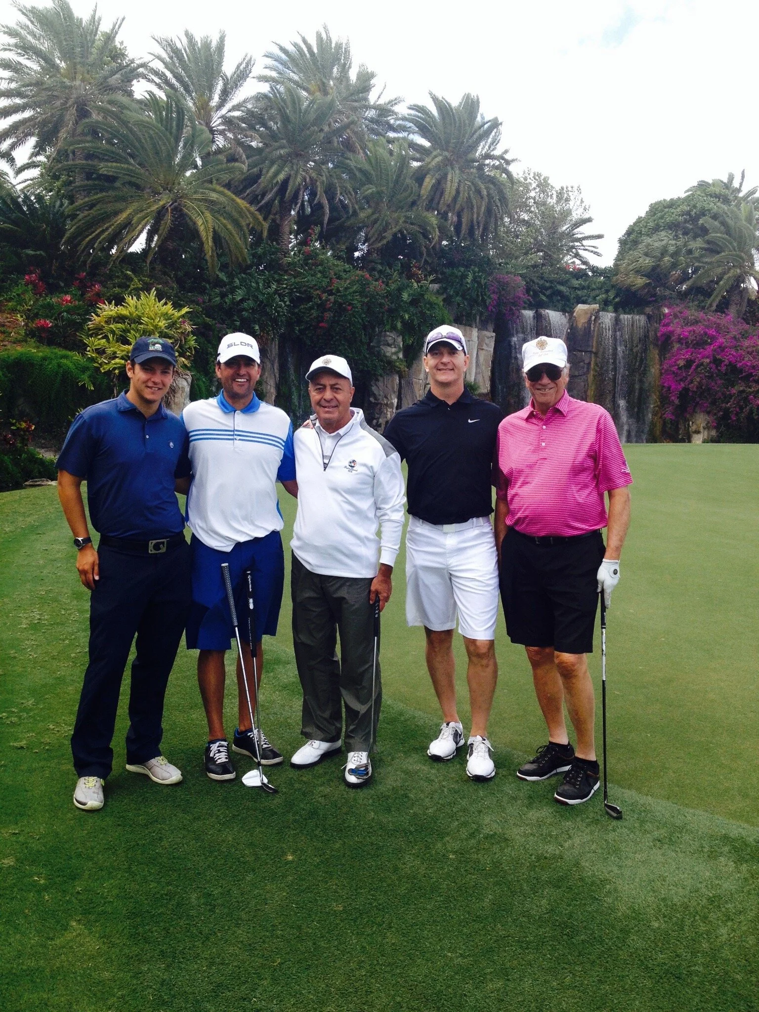 Five men standing on a golf course in front of a waterfall and tropical plants, posing for a photo with golf clubs.