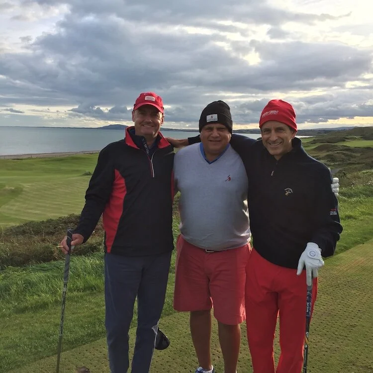 Three men standing together on a golf course by the water, smiling, with cloudy sky overhead.