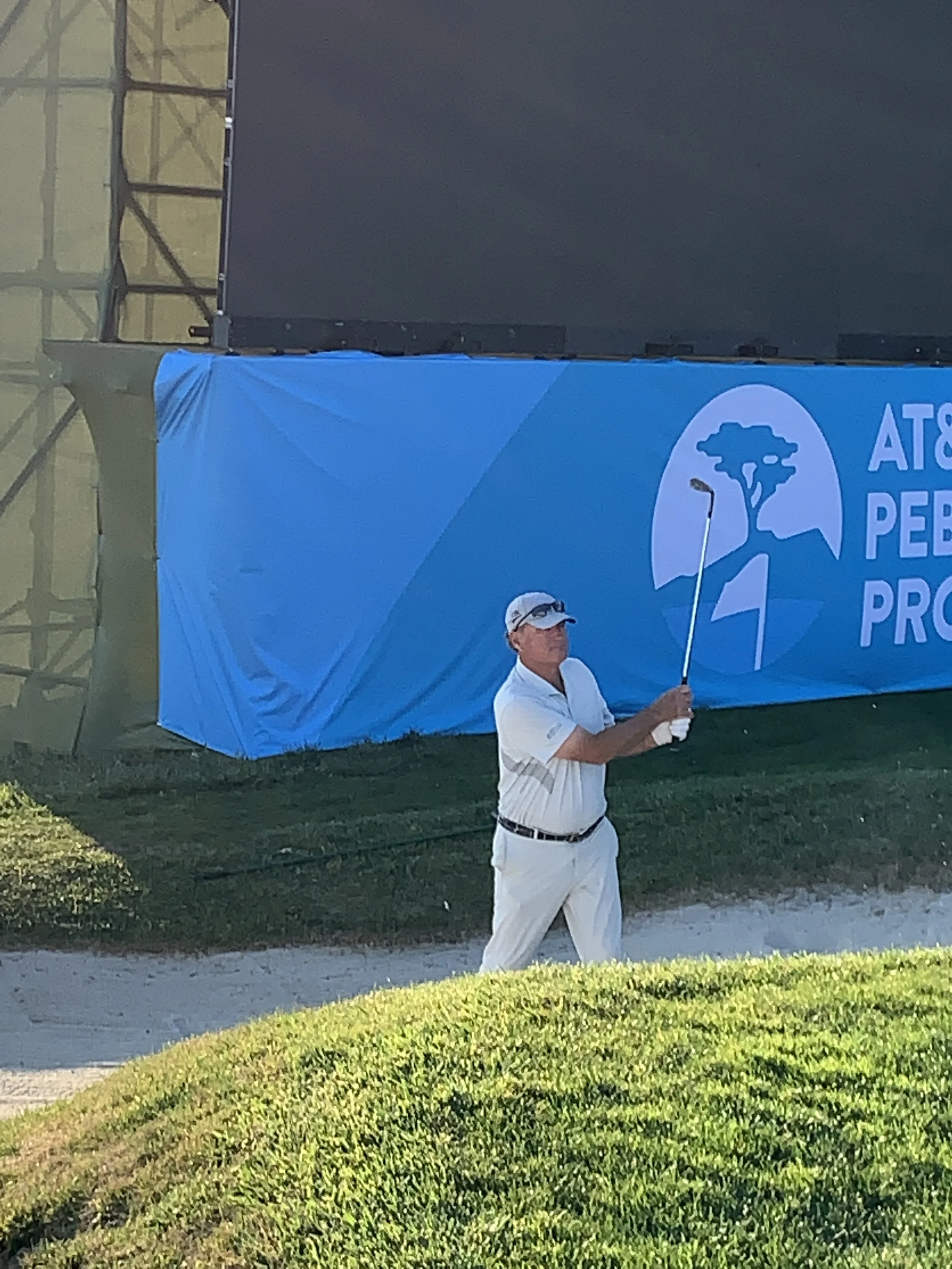 A man dressed in a white polo shirt and white pants is holding an umbrella and standing on a grassy area near a sidewalk. In the background, there is a large blue banner with white text and a logo featuring a stylized tree.