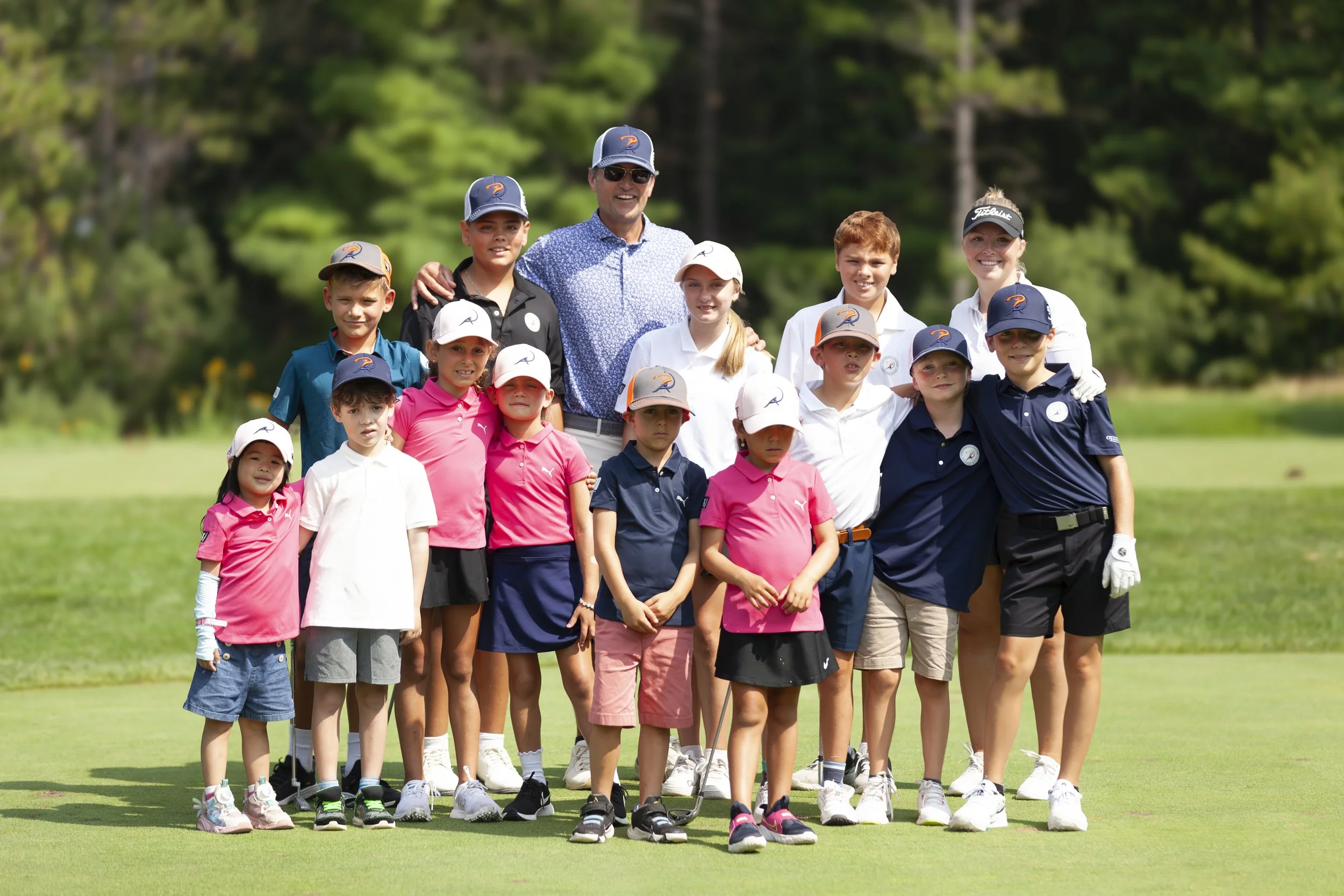 Group of children and adults standing on a golf course, dressed in golf attire, posing for a photo with lush green trees in the background.