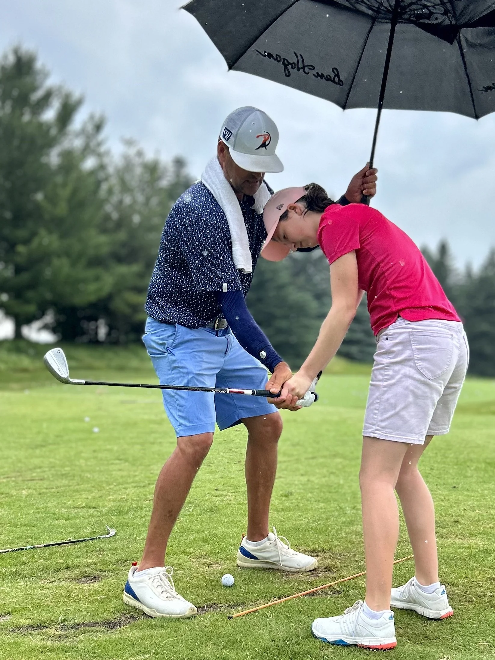 A man and a young woman practice golf on a rainy day. The man is wearing a white cap with a logo, a navy and white polka dot shirt, blue shorts, and white golf shoes. The young woman is dressed in a pink shirt, white shorts, and white golf shoes. The