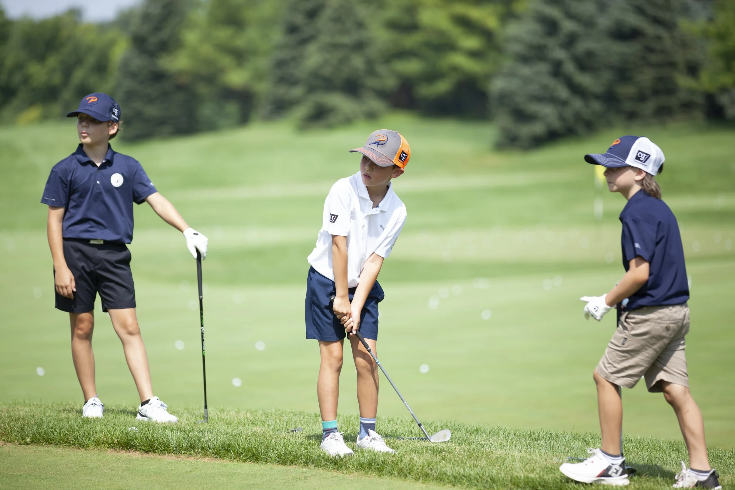 Three children playing golf on a green field, with two boys and a girl wearing hats and casual golf attire.