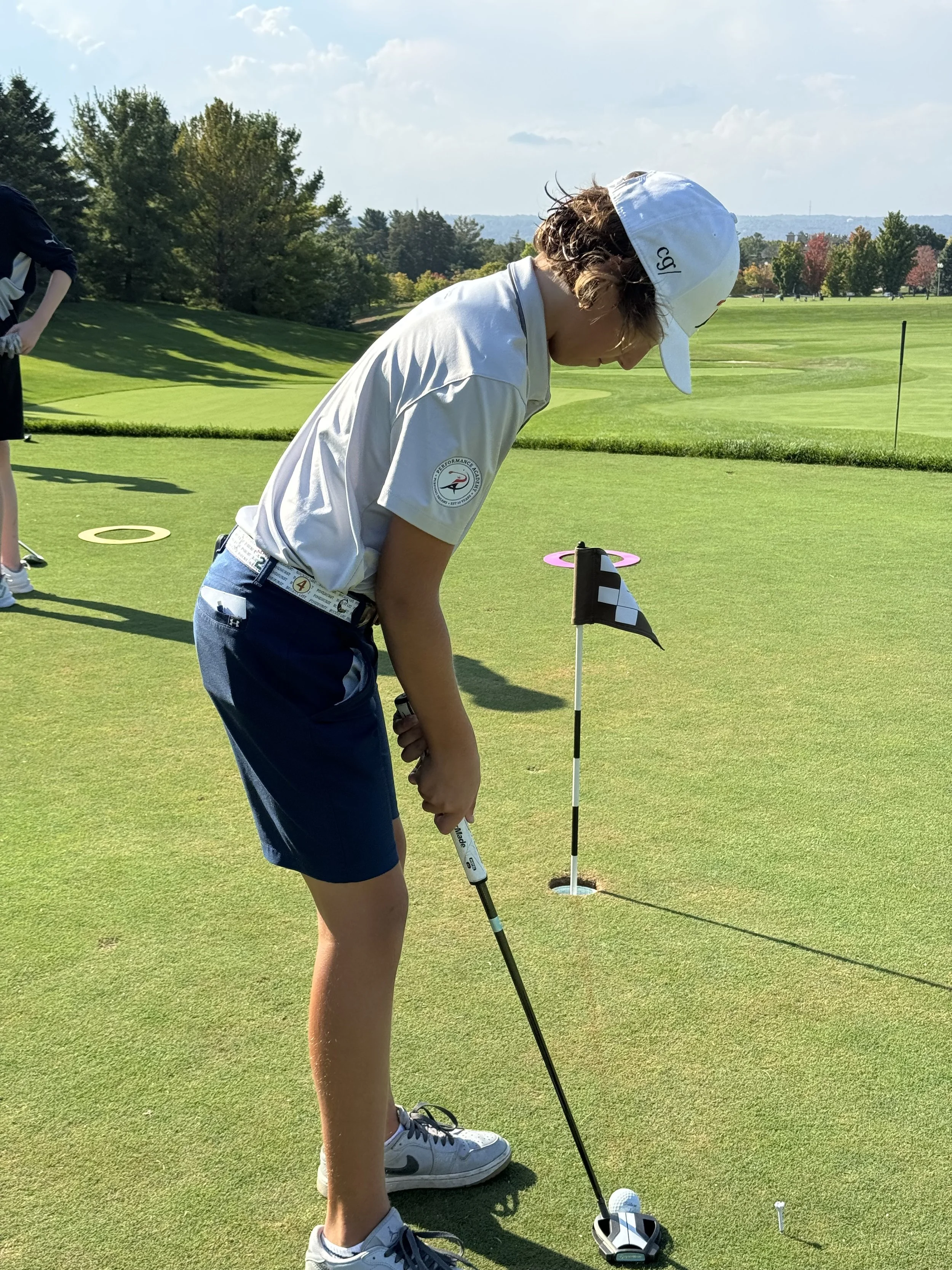 Girl in white cap and white polo shirt practicing golf on green course, with golf club and flag in foreground, trees and partly cloudy sky in background.