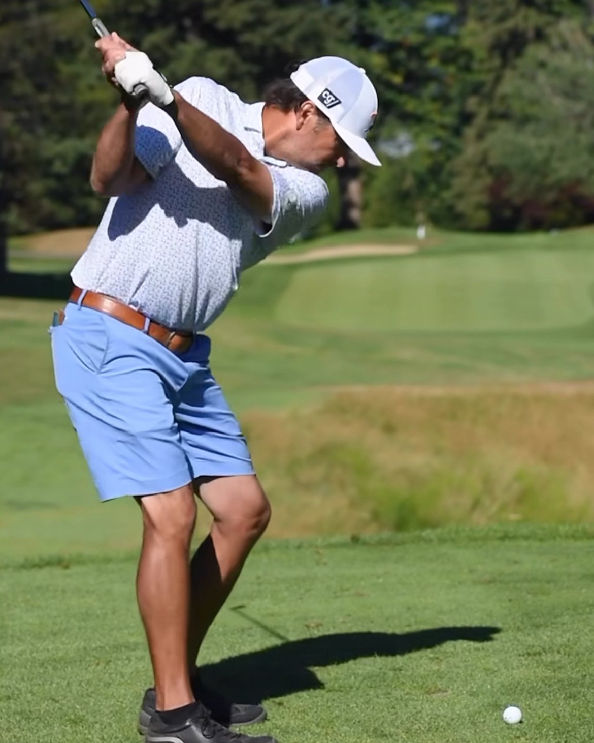 A man in a white cap, short-sleeve patterned shirt, and light blue shorts swinging a golf club on a golf course.