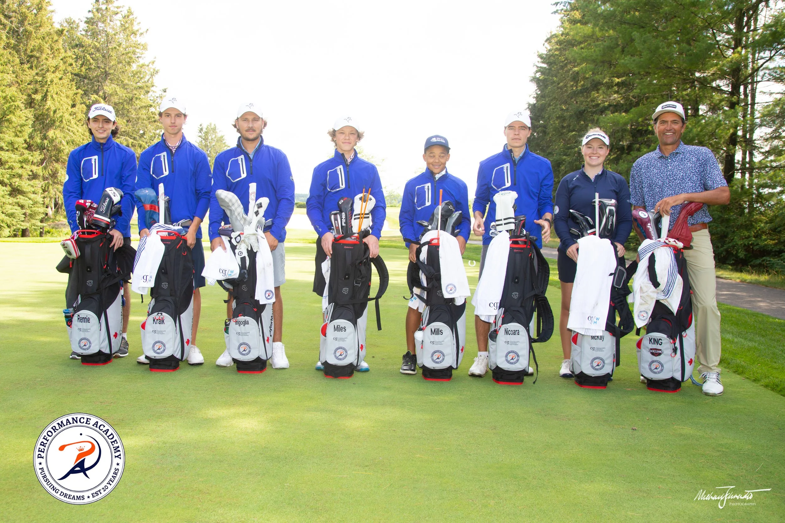 Group of young golfers and an adult standing on a golf course, each with golf bags and equipment, outdoors with trees in the background.