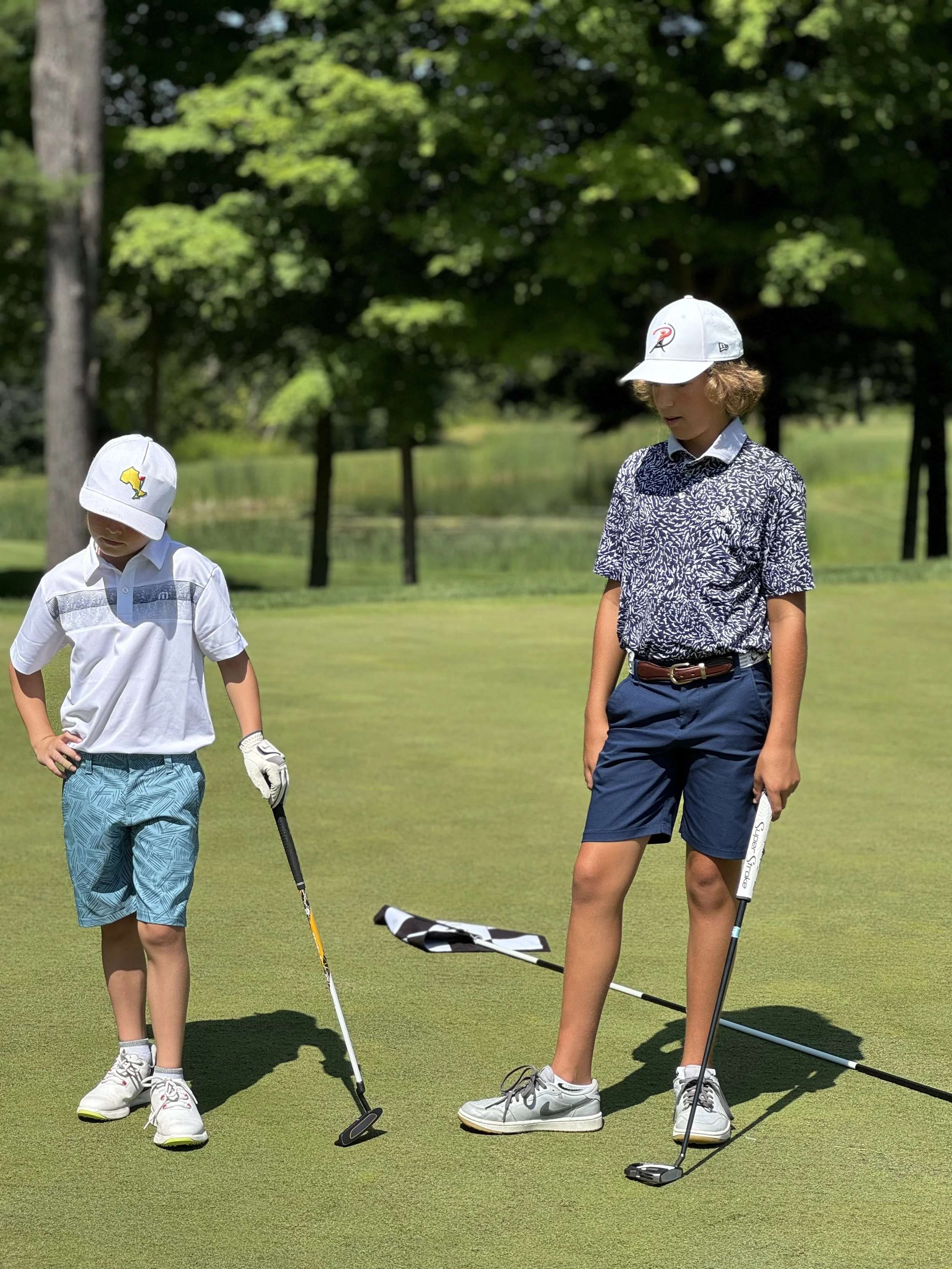 Two children playing golf on a green golf course with trees in the background. The child on the left is wearing a white hat, white shirt, and blue shorts. The child on the right is wearing a white cap, patterned shirt, and navy shorts.