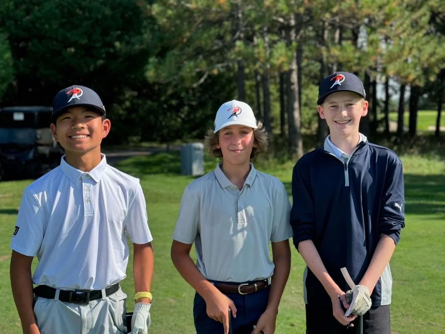 Three boys standing on a golf course, smiling at the camera, wearing golf attire and caps with a logo, surrounded by trees.