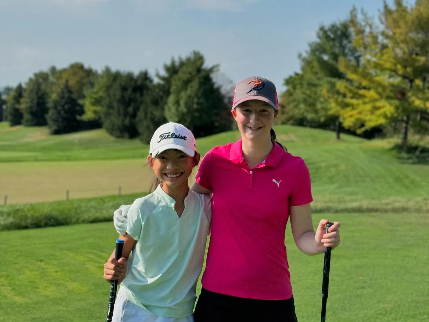 Two young girls standing on a golf course holding golf clubs, smiling at the camera. One girl wears a white cap and light-colored shirt, the other wears a pink shirt and a gray cap. Trees and grass are visible in the background.