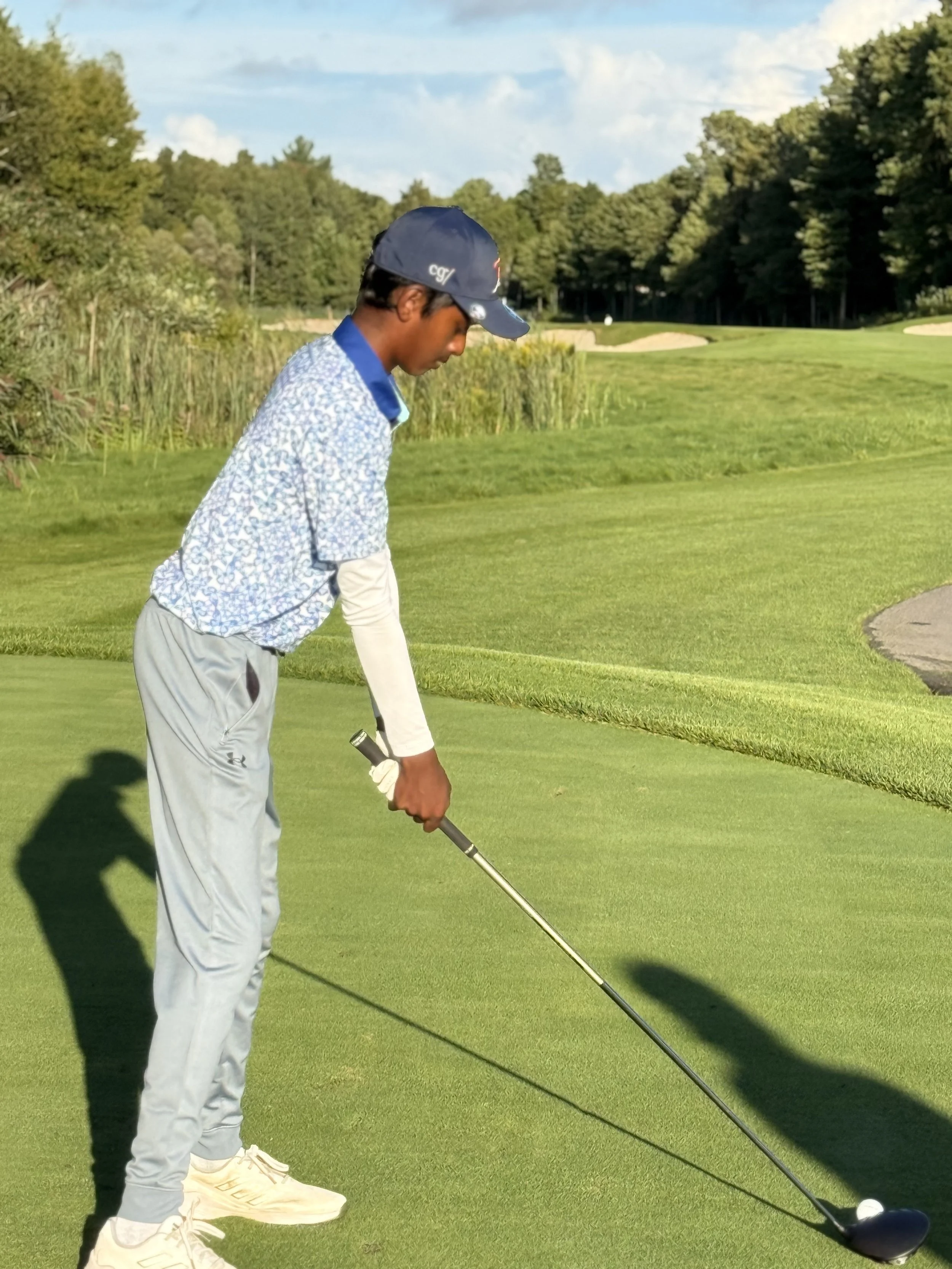 A young male golfer wearing a blue cap, patterned shirt, and gray pants on a golf course, preparing to hit a golf ball with a driver club, with trees and sand bunkers in the background.