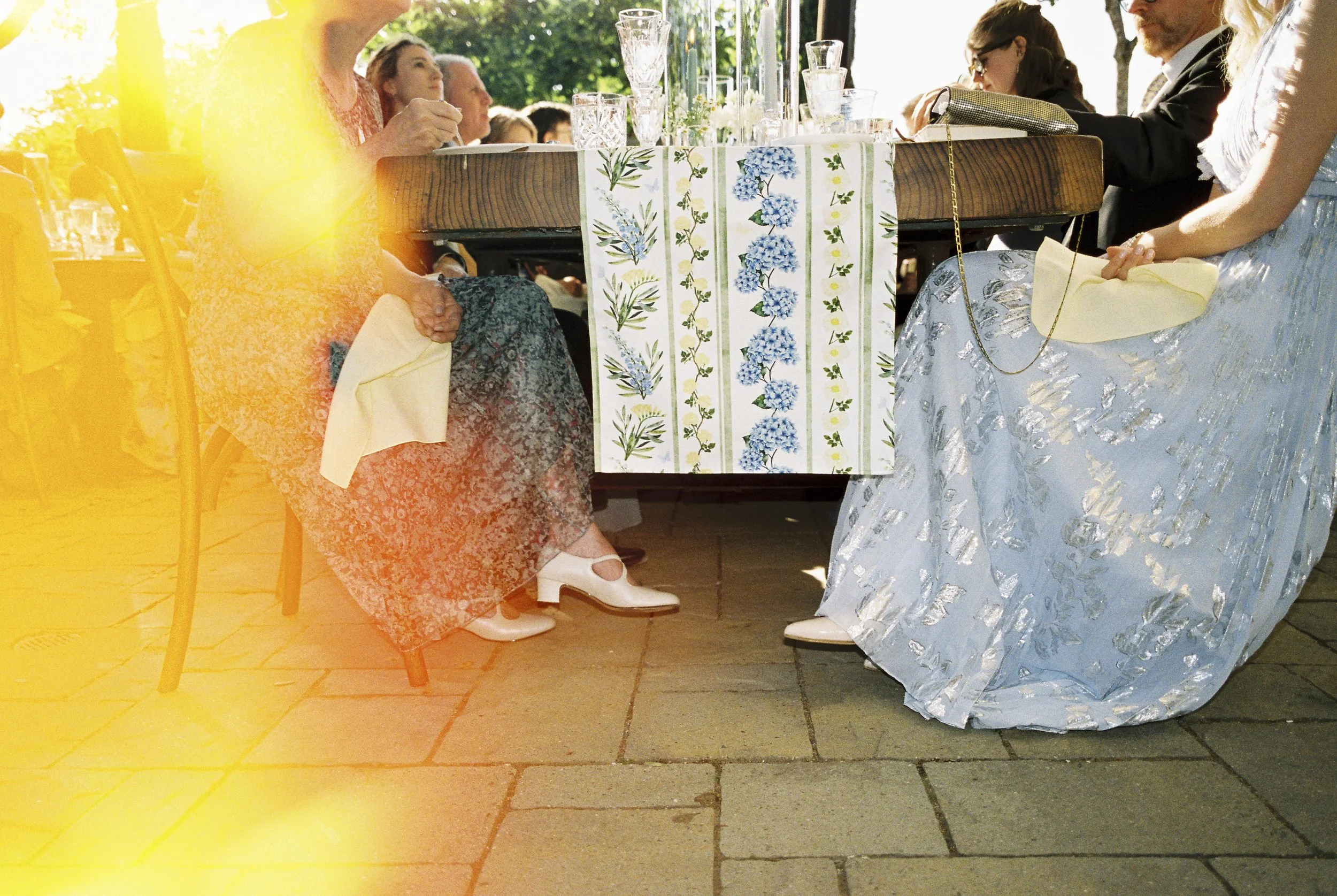 People sitting around a table at an outdoor event, with a floral tablecloth and glasses on the table, wearing formal dresses and suits, with sunlight filtering through trees.