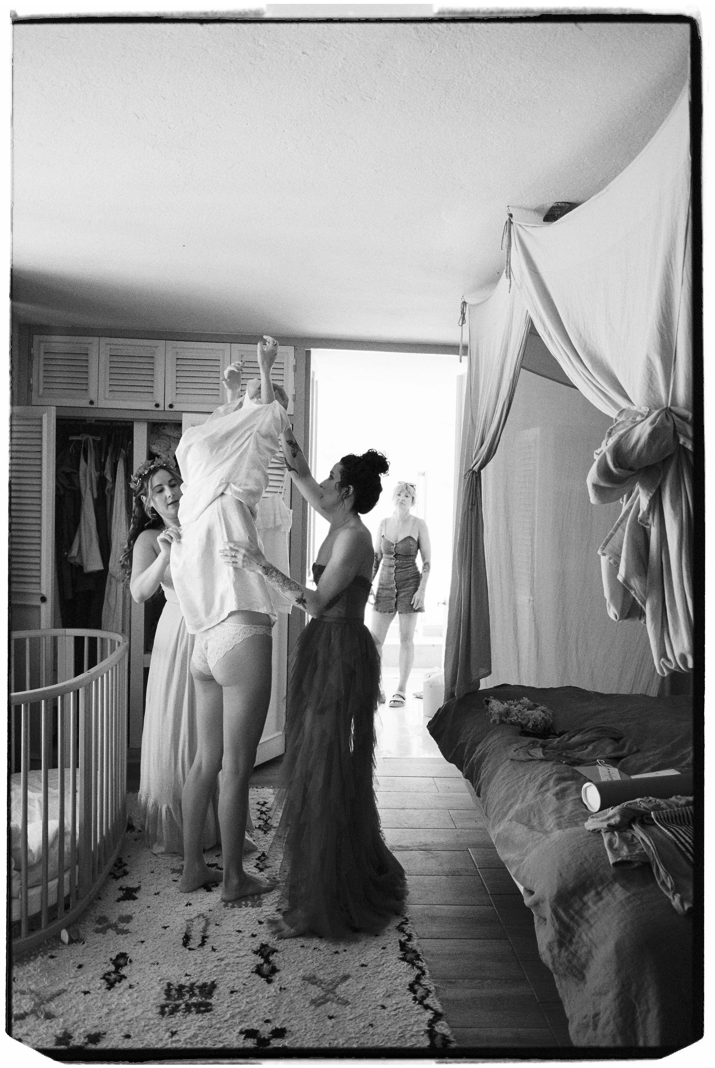 Four women in a bedroom, one being helped with her dress, with a crib and a bed in the room, and a woman in shorts and a tank top standing near the doorway.