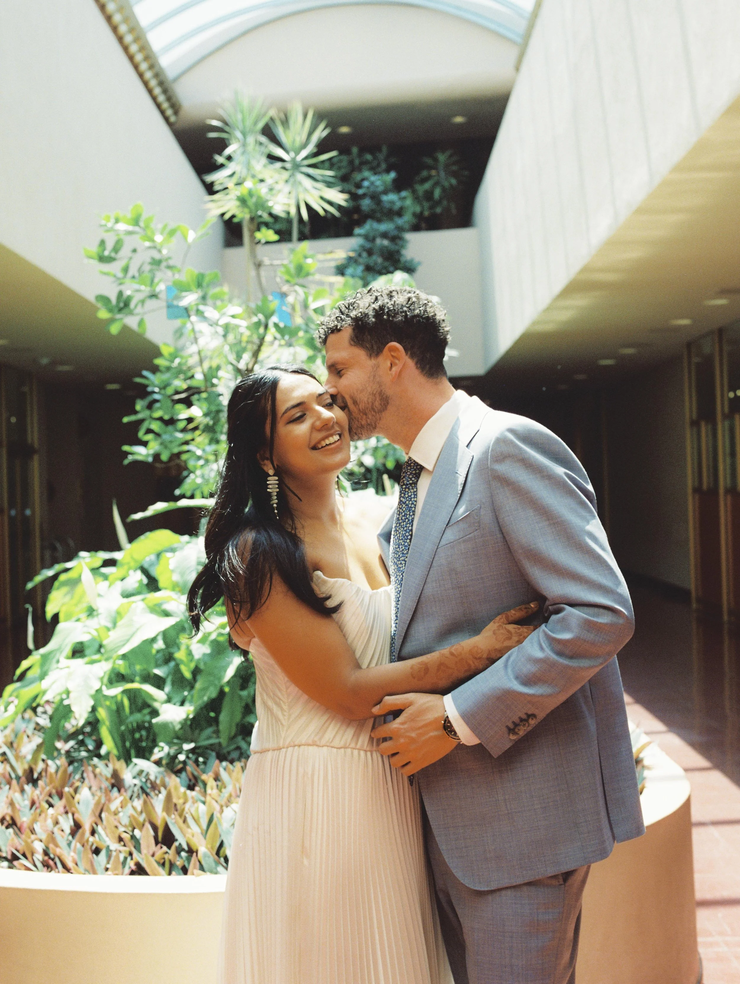 A happy couple is embracing and sharing a kiss in a brightly lit indoor space with lush greenery and plants around them.