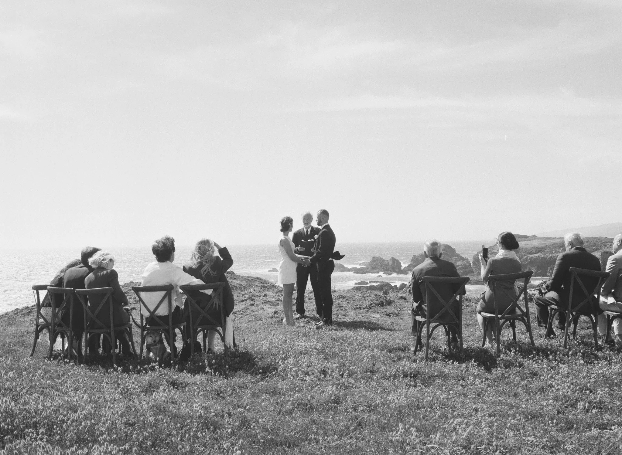 A black-and-white photo of a seaside wedding ceremony with guests seated on chairs on grass, and the officiant wedding couple in the center, holding hands, with ocean and cliffs in the background.