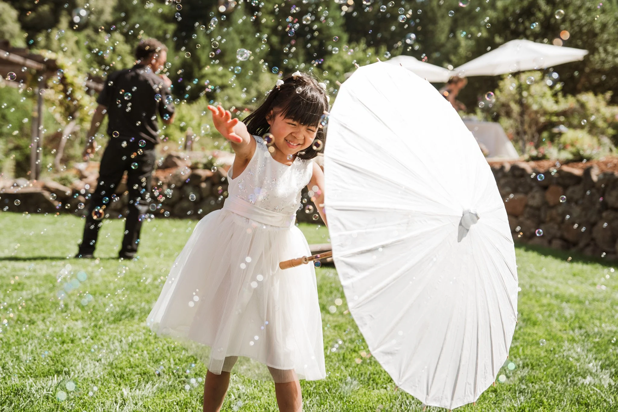 A young girl in a white dress playing with a white parasol and bubbles outdoors in a sunny garden, with an adult man and white umbrellas in the background.