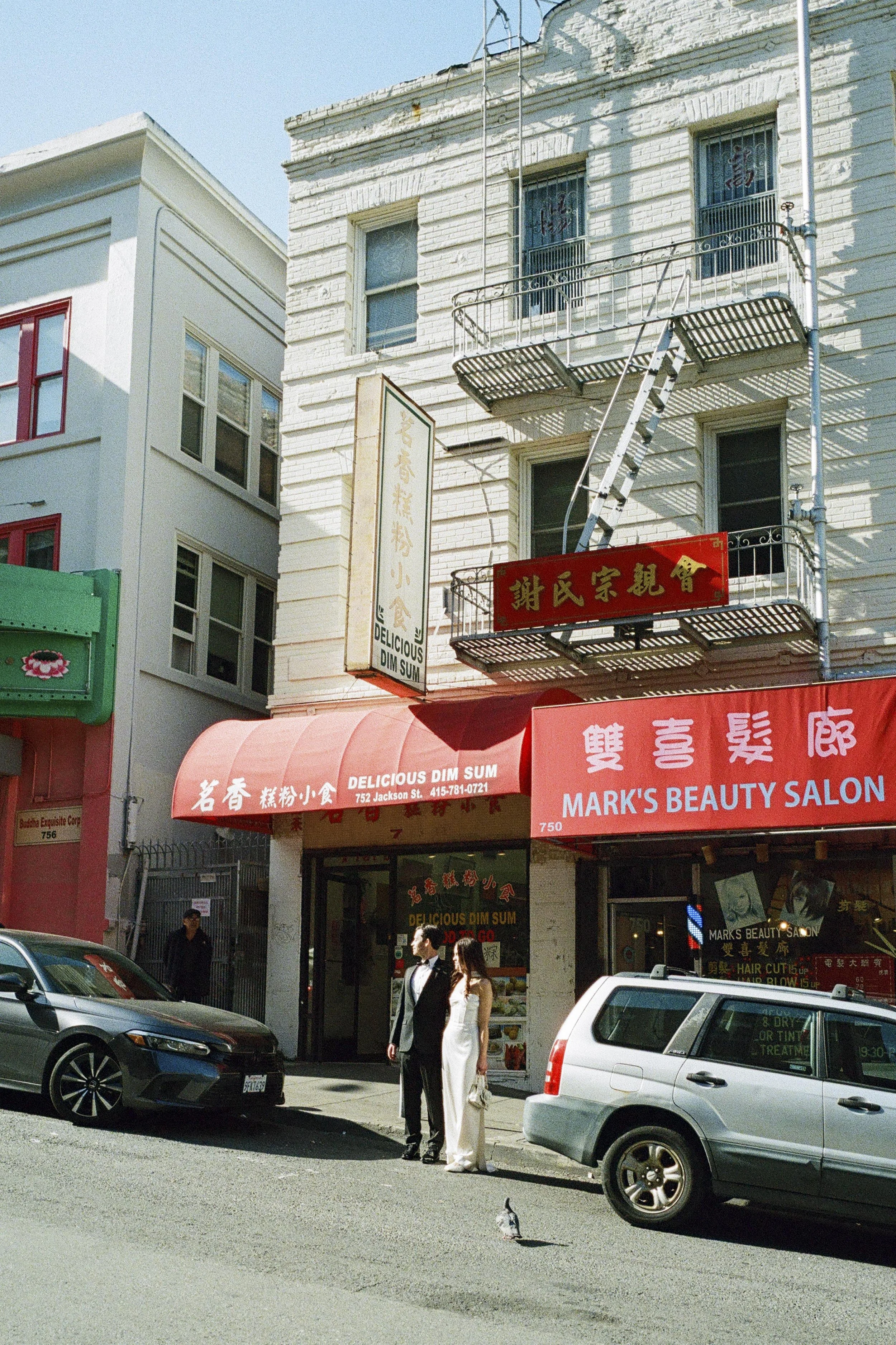 A street scene in front of a restaurant called 'Delicious Dim Sum' and a beauty salon called 'Mark's Beauty Salon.' There are two people dressed in formal attire standing on the sidewalk and two cars parked nearby. The buildings have signs in Chinese