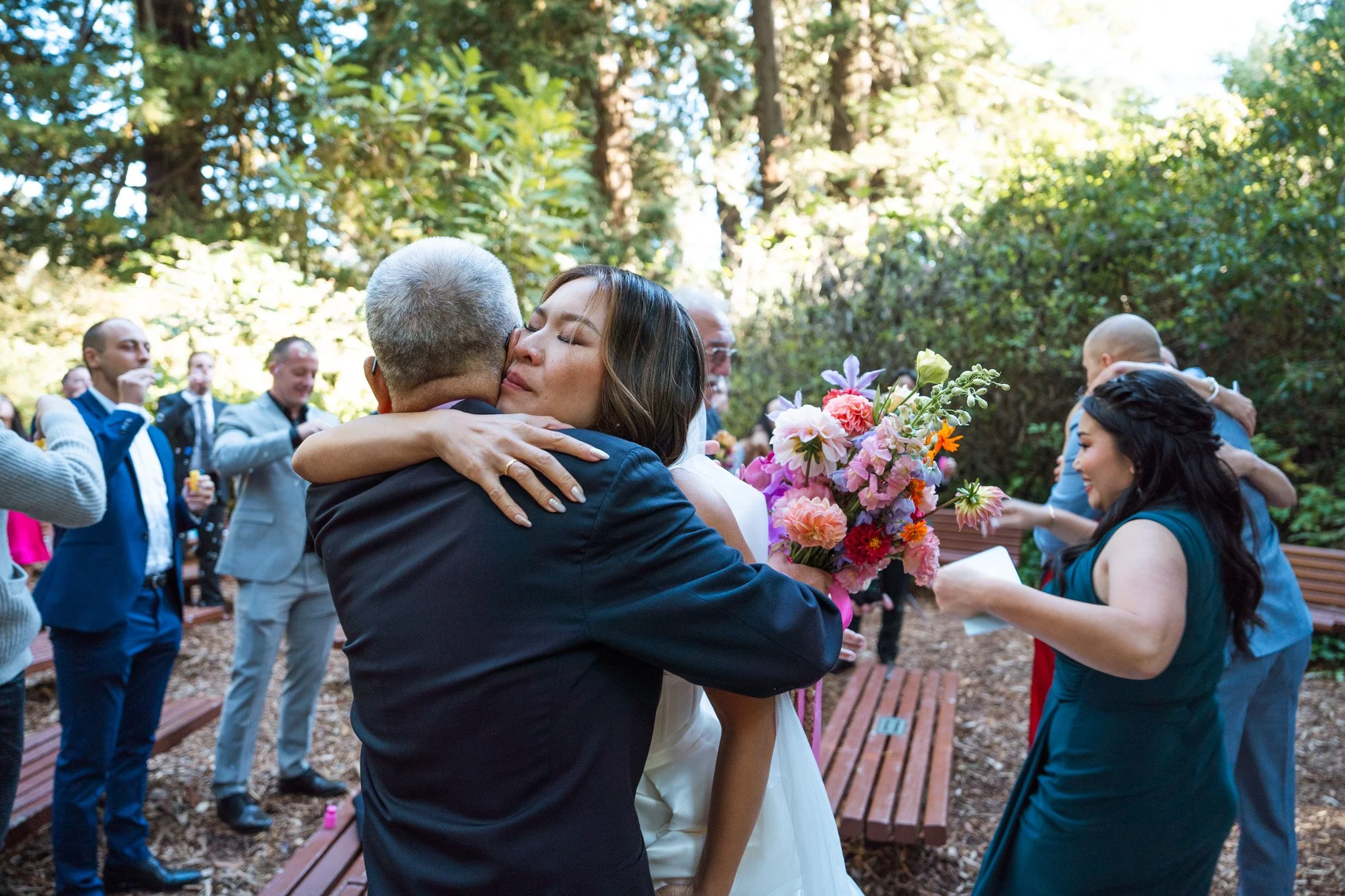 A woman in a white dress hugging a man in a dark suit, holding a colorful bouquet of flowers, at an outdoor celebration with guests embracing in the background.