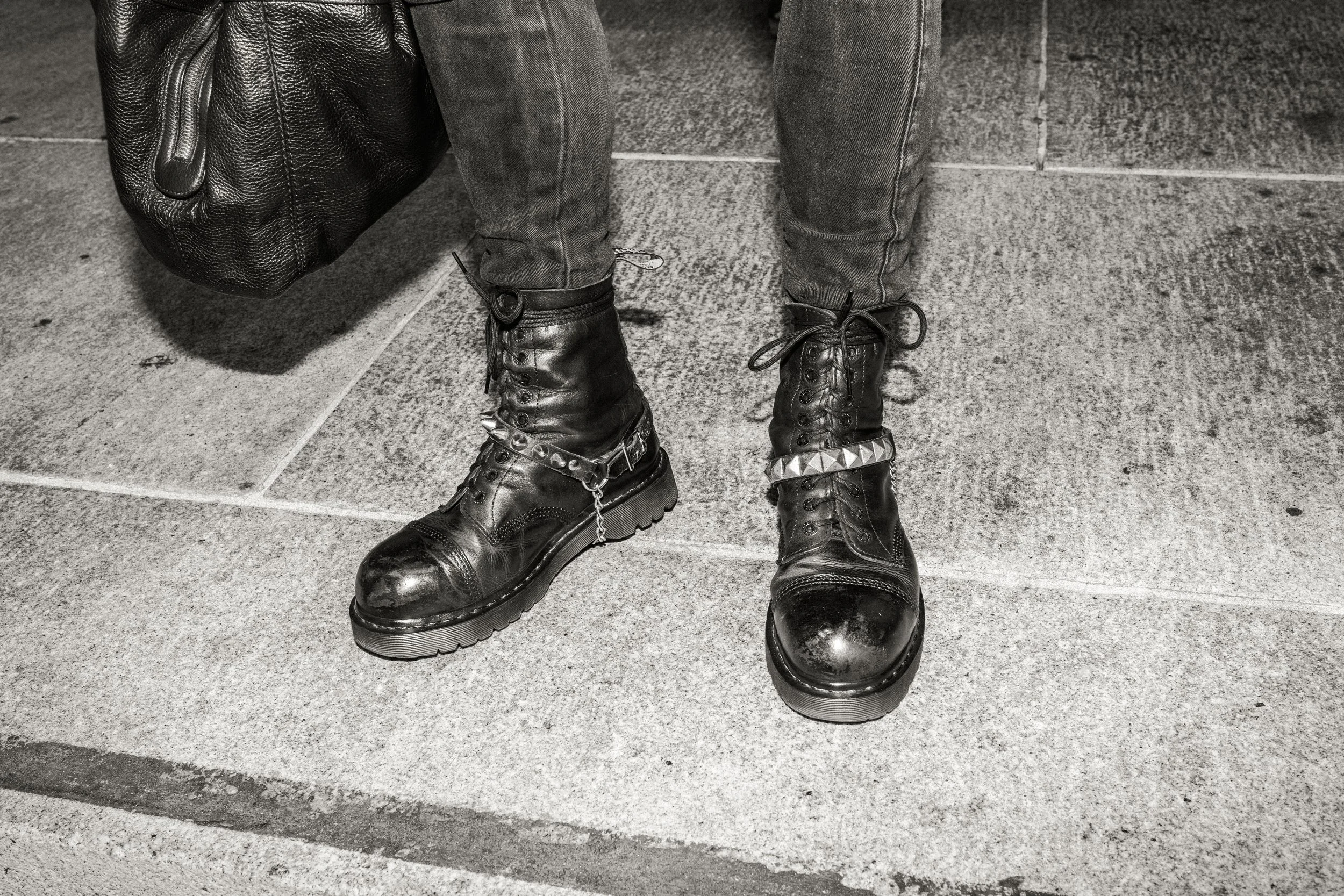 Close-up of person wearing black leather boots with metal embellishments and dark jeans, standing on a city sidewalk, carrying a black textured bag.