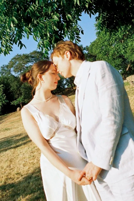 A couple dressed in white holding hands and touching foreheads in a park with green trees and blue sky.