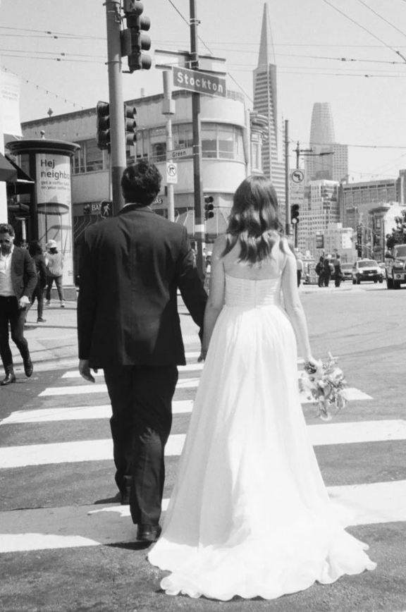 A bride and groom crossing a city street, with the bride holding a bouquet of flowers, in downtown San Francisco, with skyscrapers in the background, black and white photo.