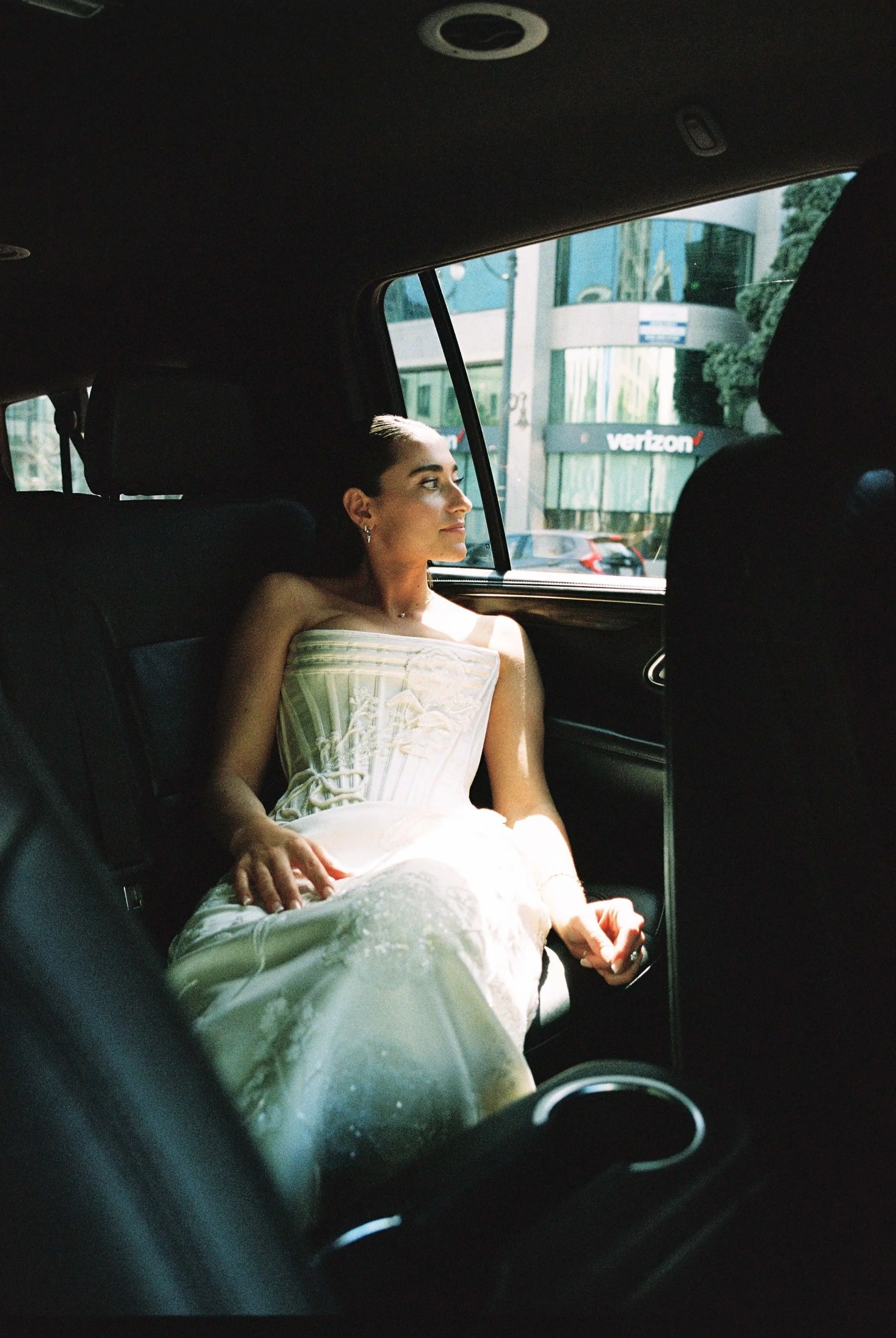 A woman in a wedding dress sitting in the backseat of a car, looking out the window.