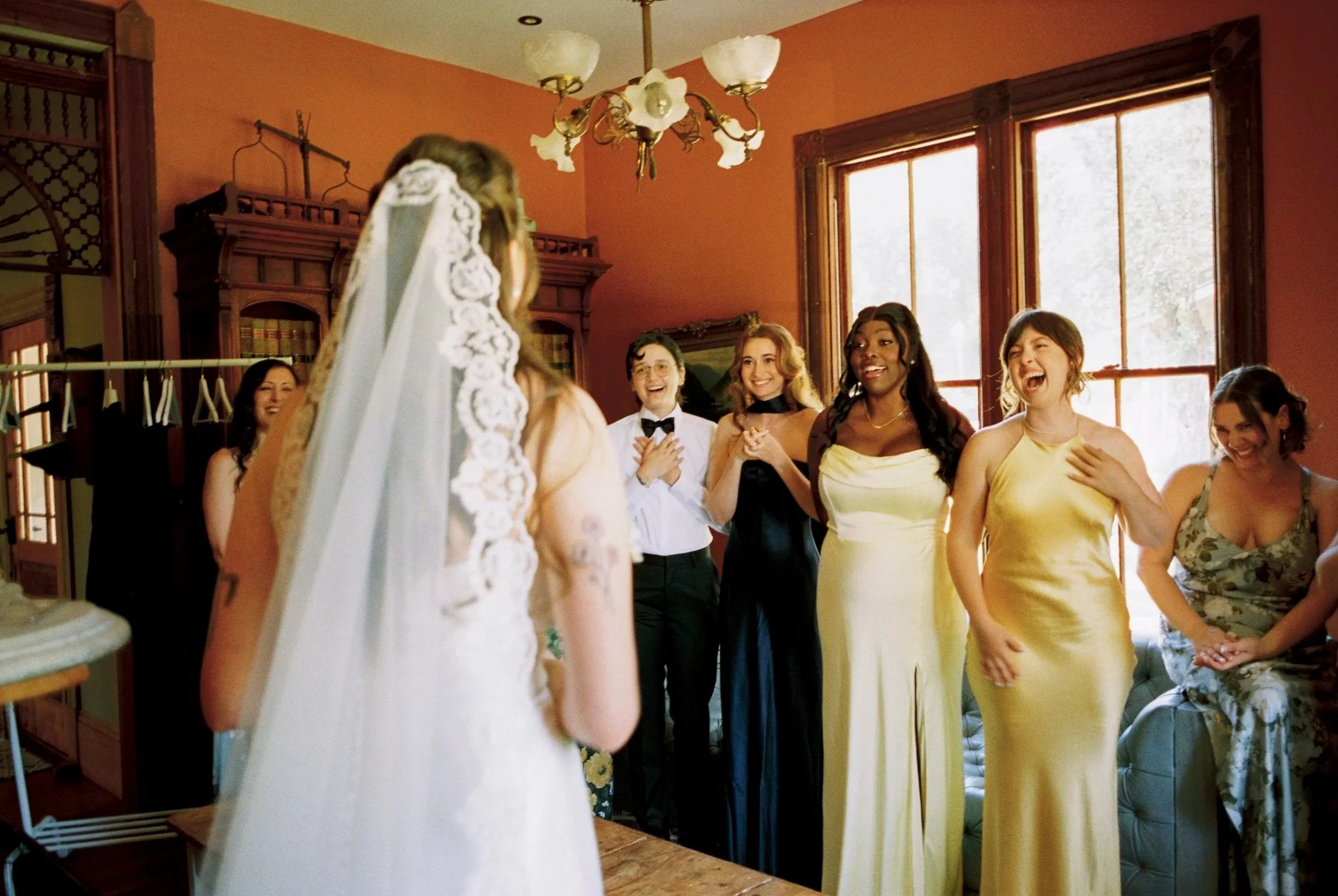 A bride dressed in white with a veil standing in front of five women and one man, all smiling and laughing, in a room with red walls, large windows, and a wooden cabinet.