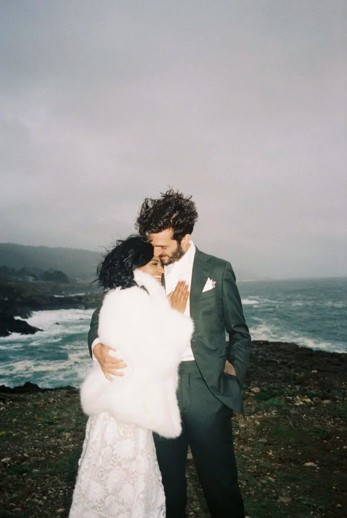 A couple dressed in wedding attire embraces on a rocky coastline during overcast weather.