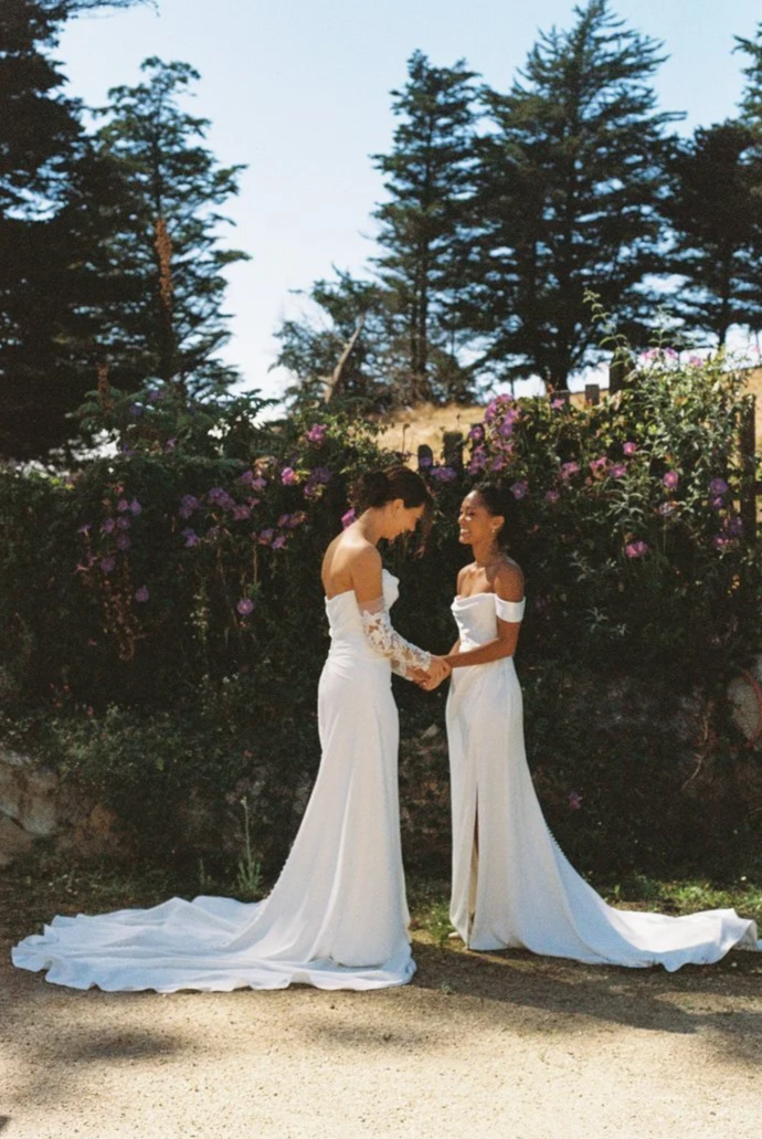 Two women dressed in white wedding dresses holding hands and smiling outdoors in front of purple flowering bushes and tall trees.