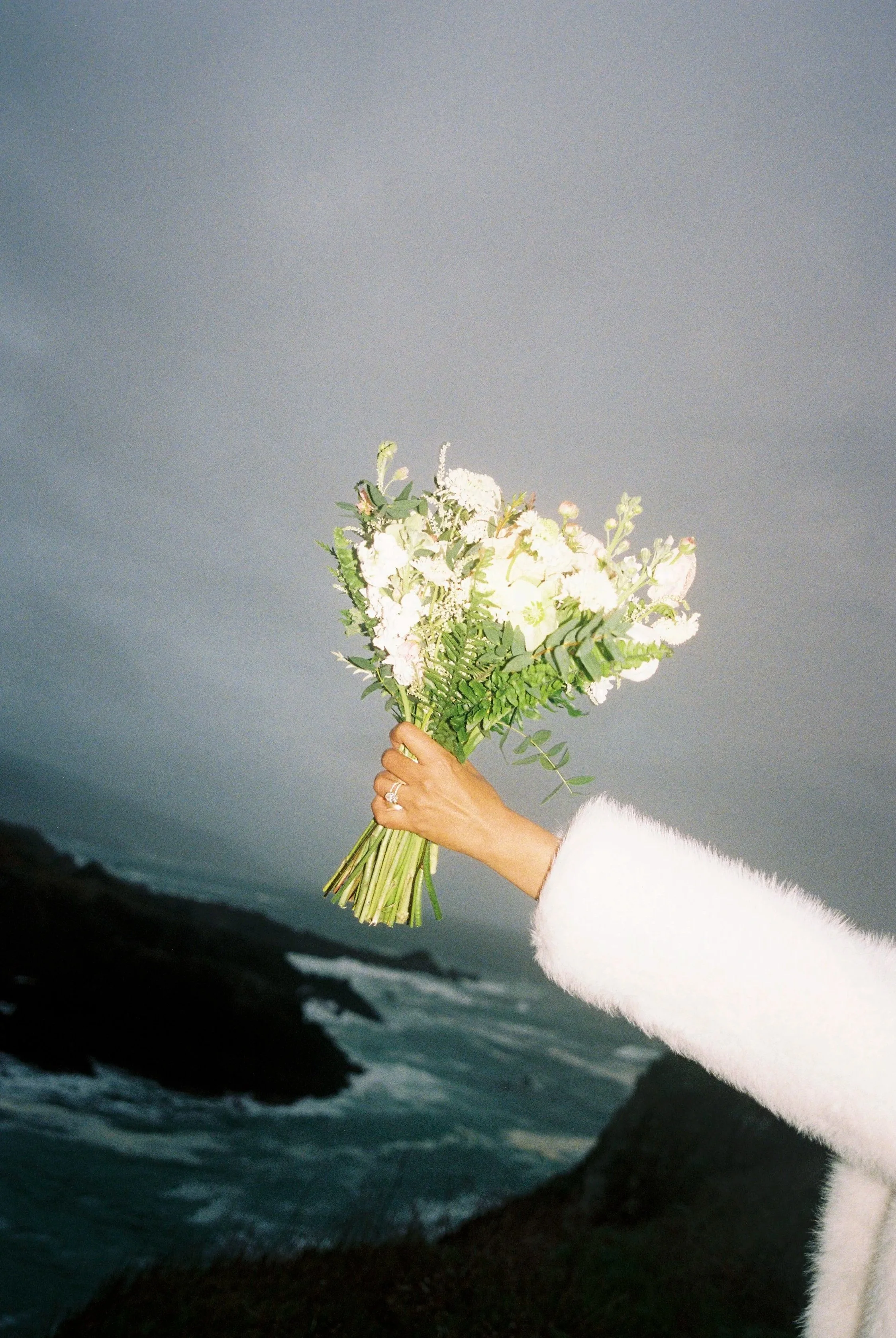 A person holding a bouquet of white flowers with an ocean and rocky coastline in the background, wearing a white fuzzy coat.