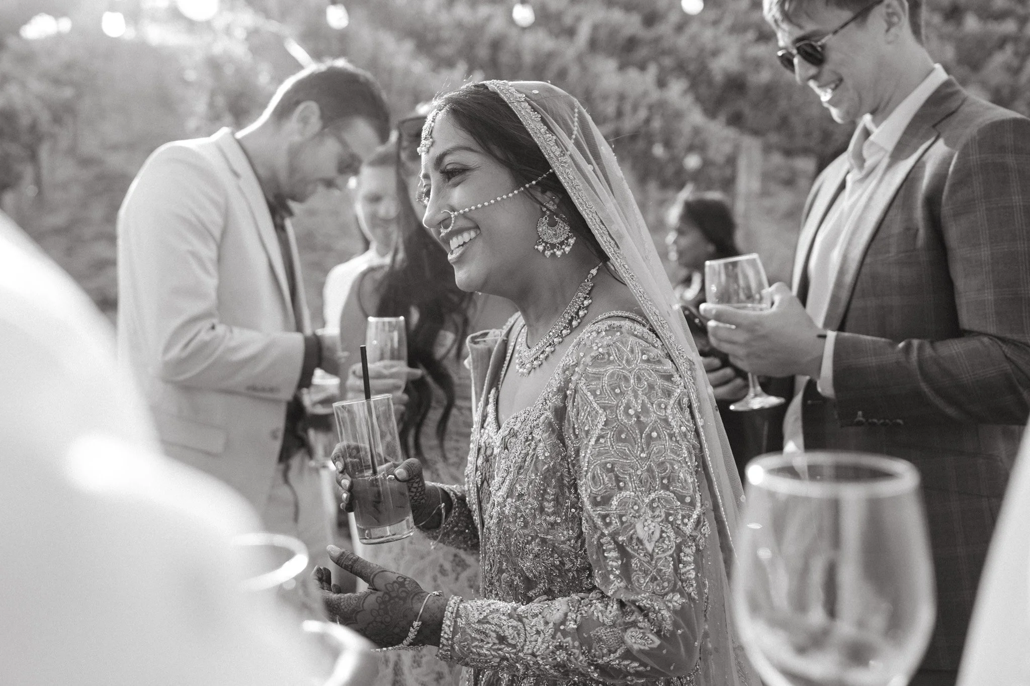 Group of people at an outdoor wedding reception, featuring a woman in traditional ornate attire with henna on her hands, holding a glass, smiling and interacting with others.