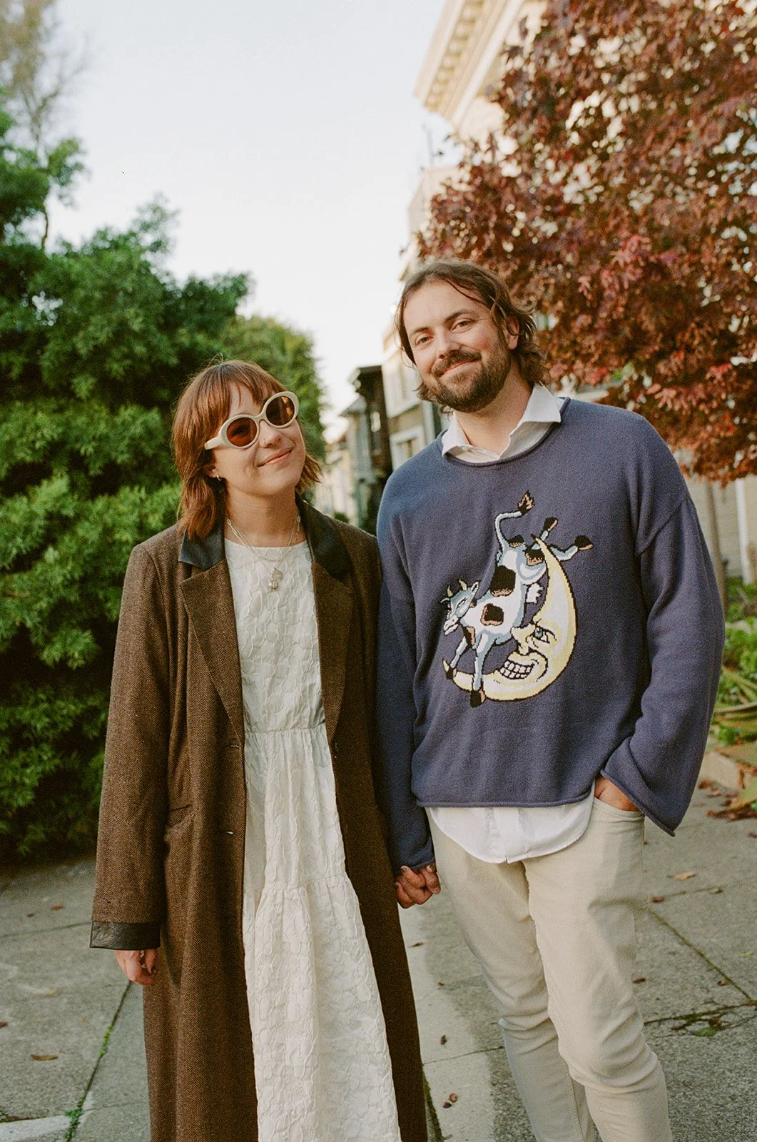 A man and woman standing outside holding hands, smiling, with trees and buildings in the background.