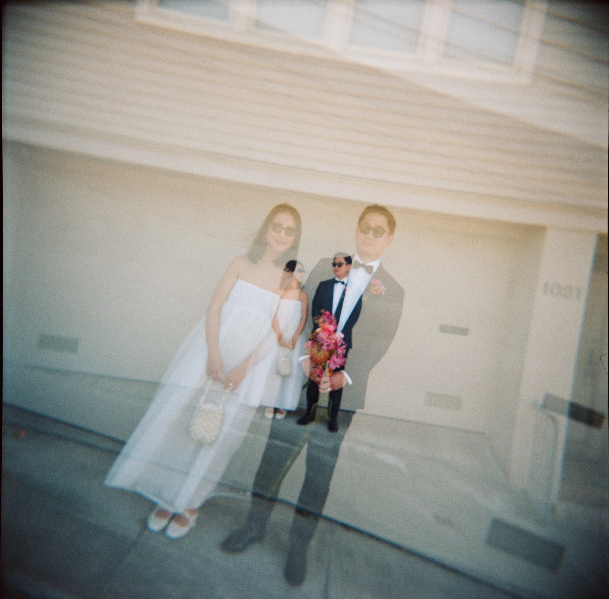 A double exposure photo of a couple in wedding attire standing in front of a house, with the man holding a bouquet of flowers and the woman in a white dress and glasses.