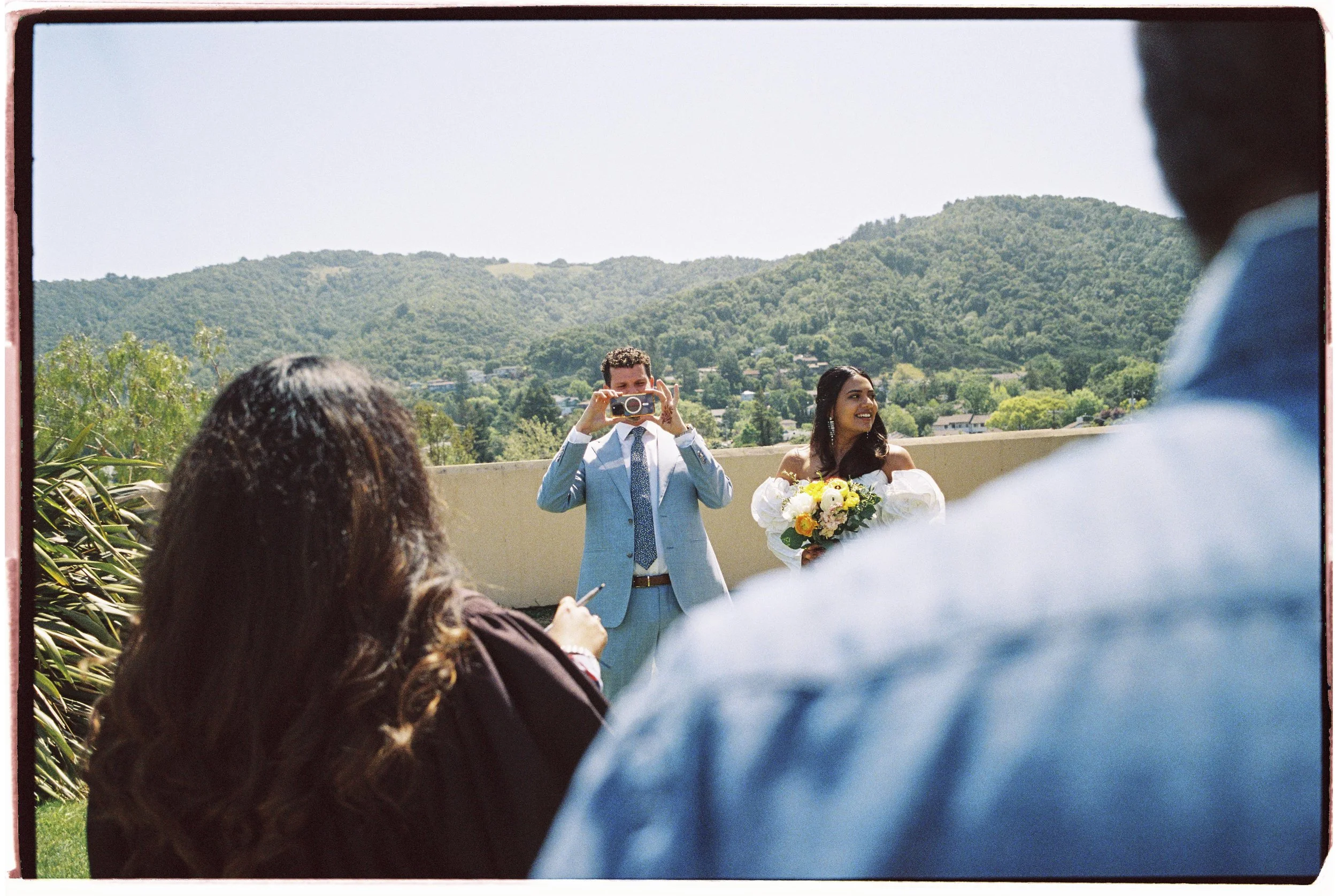 People at a wedding ceremony outdoors, with hills and trees in the background. A person is taking a photo of the bride, who is holding a bouquet and smiling. Other wedding guests are visible, with one person in the foreground using a phone.