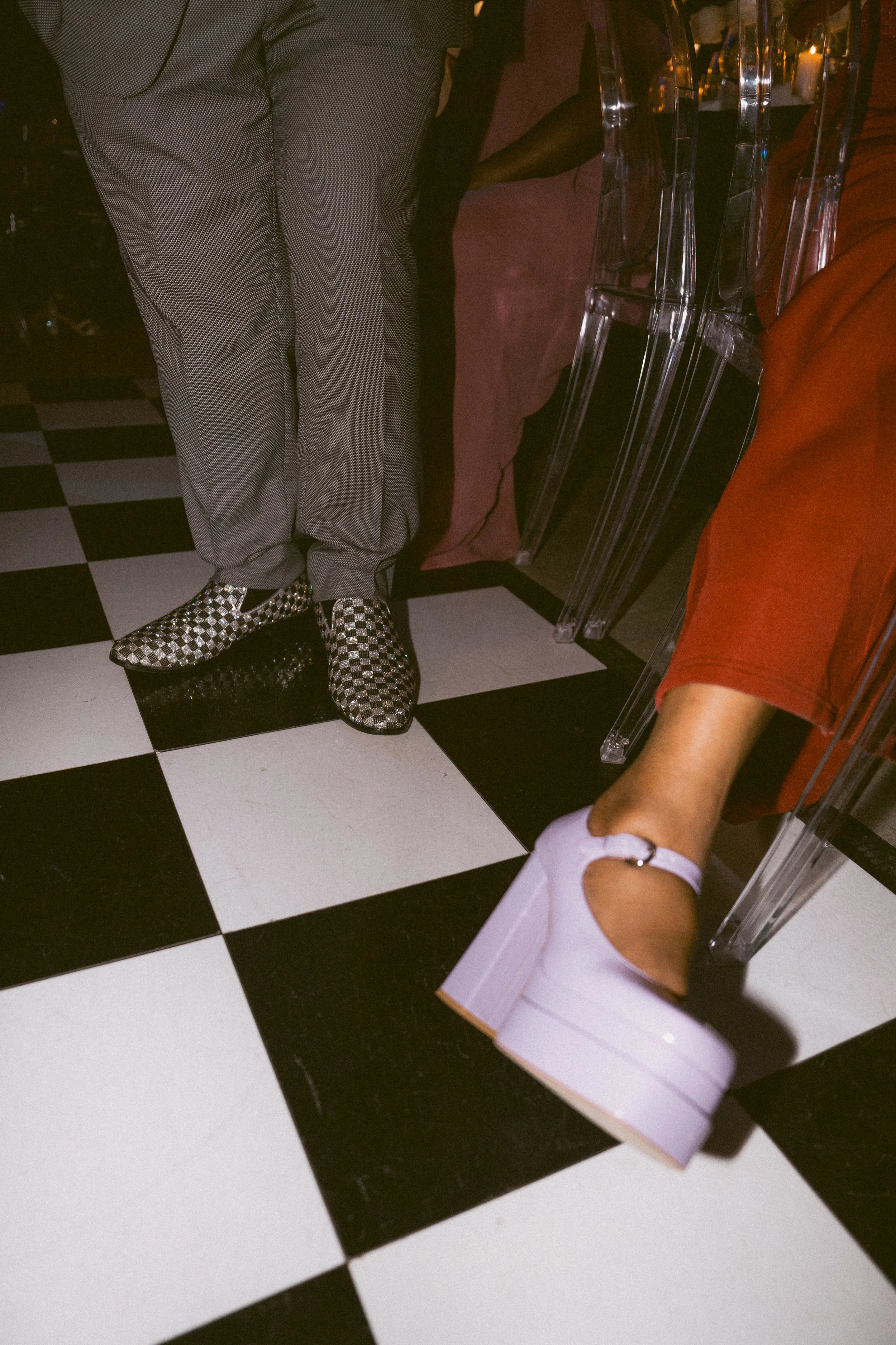 Close-up of two people sitting on clear acrylic chairs on a black-and-white checkered floor, showcasing their stylish footwear and clothing.