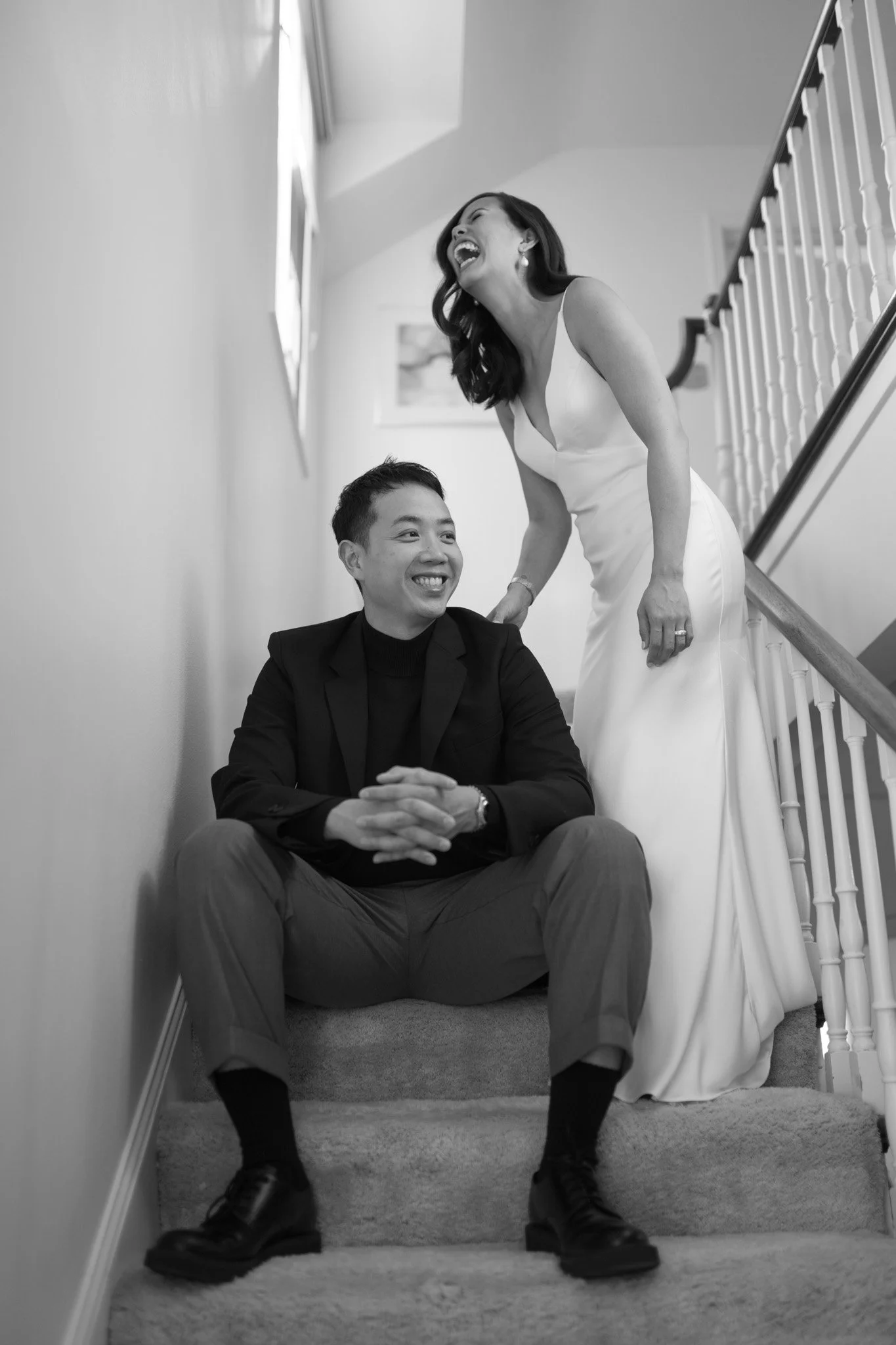 A black-and-white photo of a woman in a wedding dress and a man in a suit sitting on stairs, laughing and smiling together.