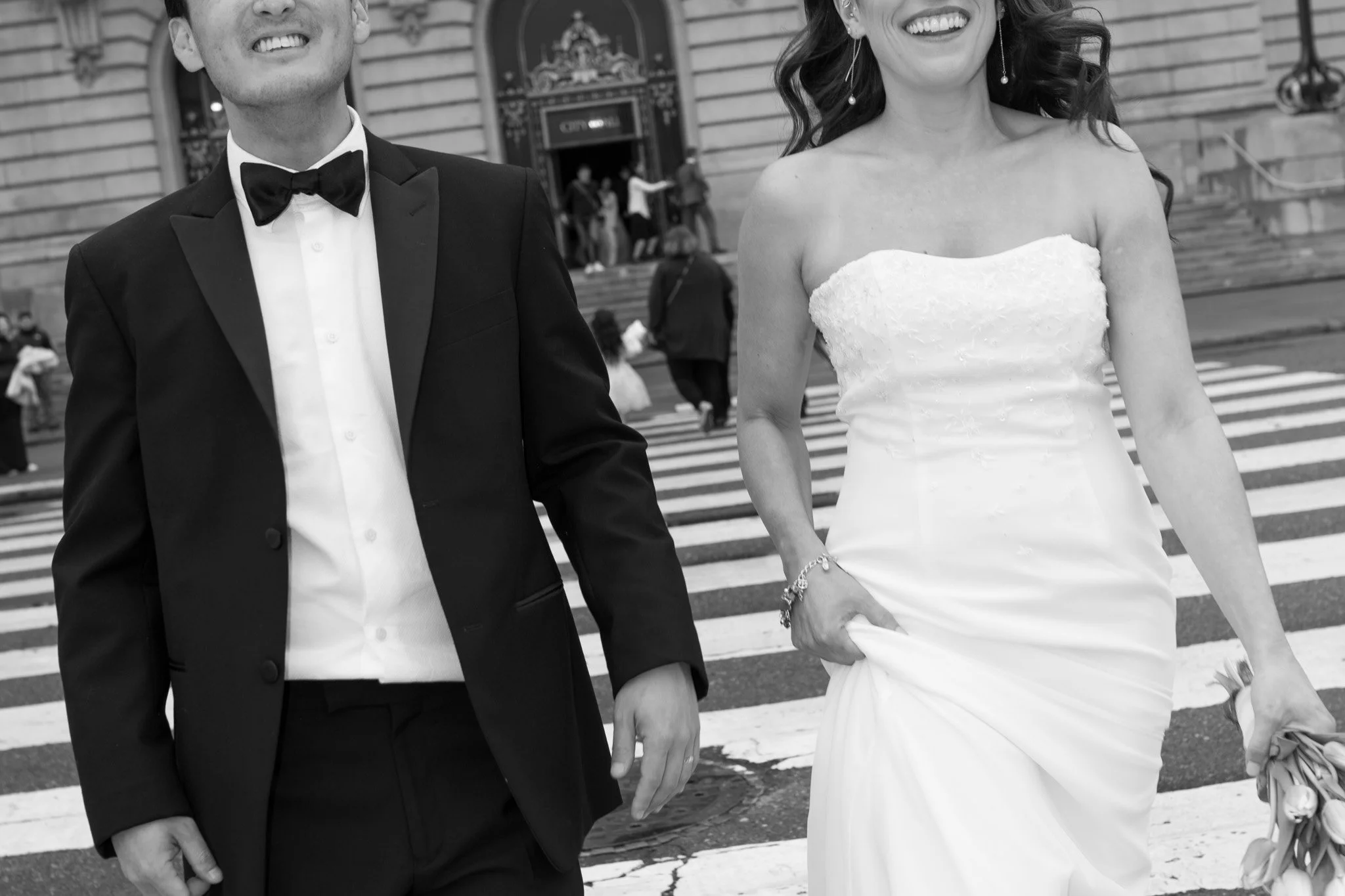 Bride and groom smiling and walking in a crosswalk, wearing wedding attire.
