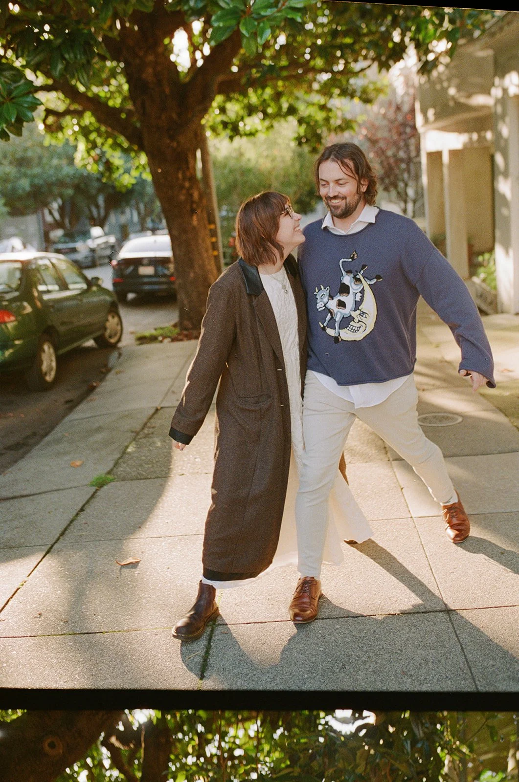 A man and woman walking on a sidewalk under a large tree, smiling, with parked cars in the background during daytime.