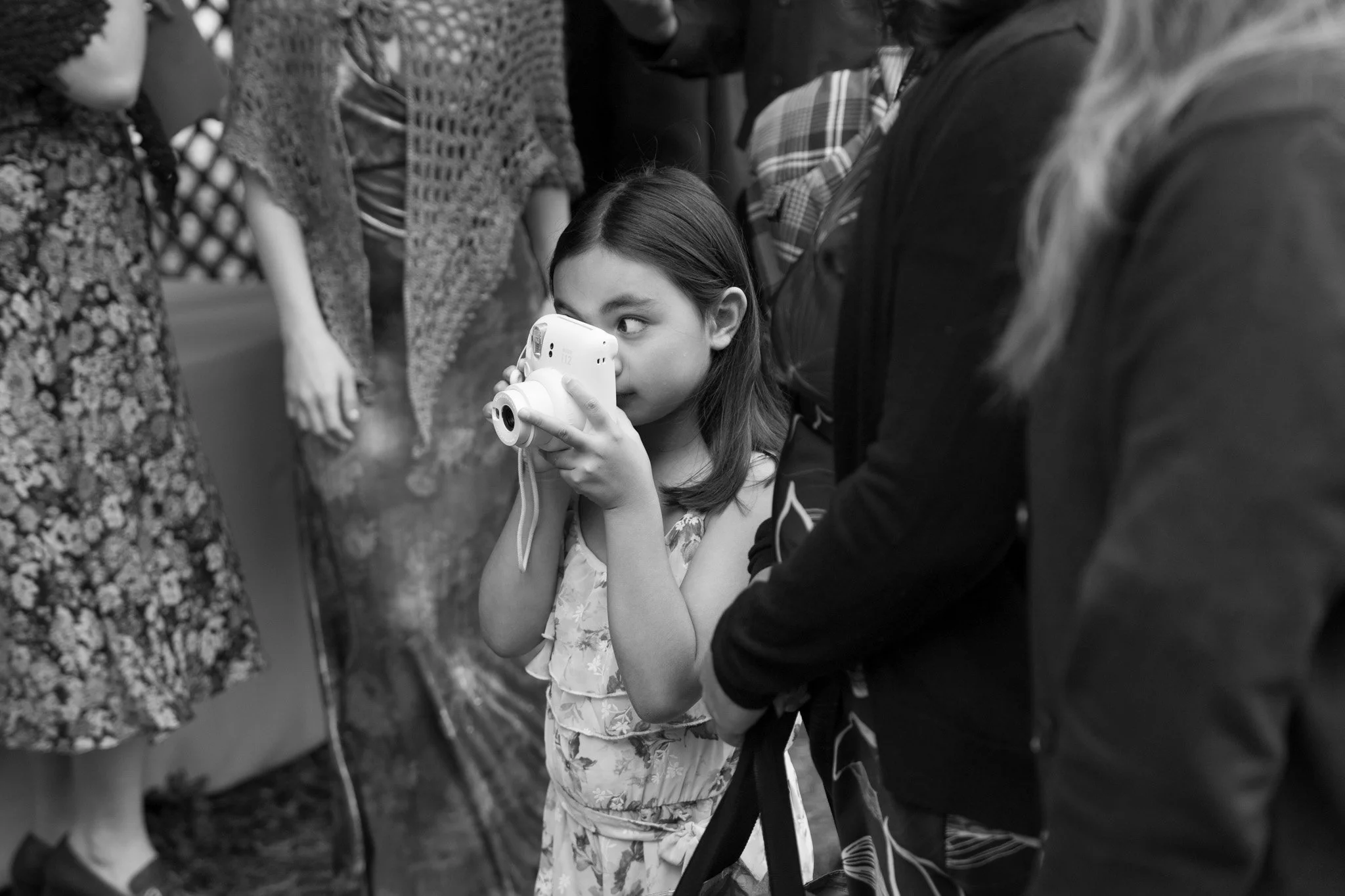 A young girl with dark hair holding a camera, surrounded by people dressed in various floral and patterned clothing, apparent at an indoor event.