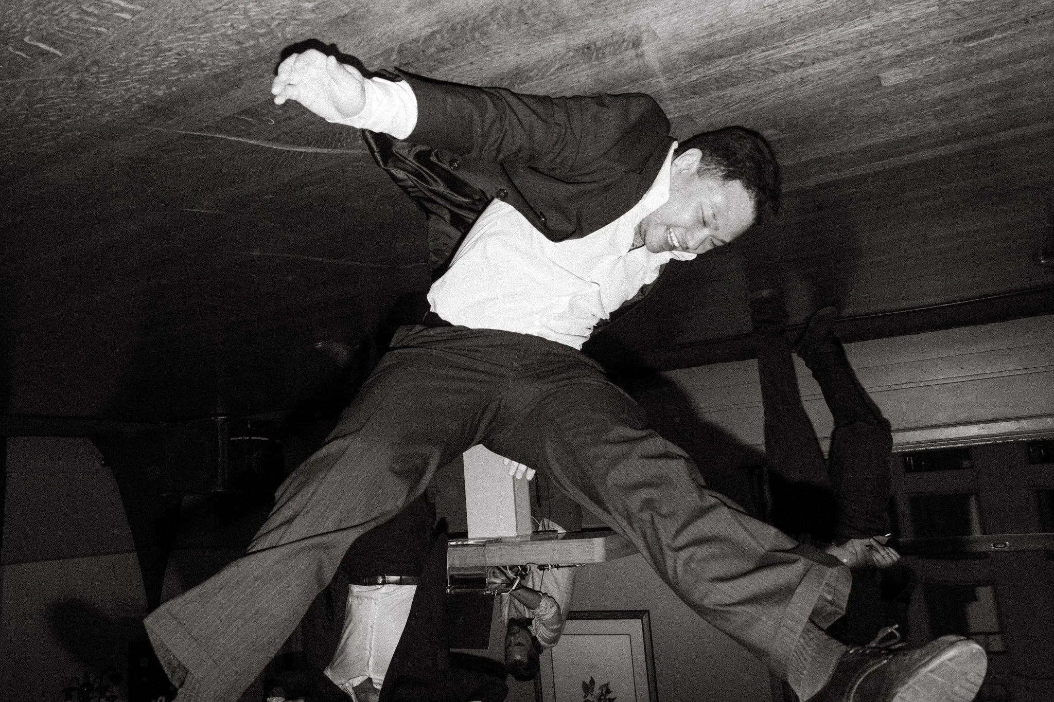 Person in midair wearing a suit, appearing to be jumping or dancing in a black and white photo, with an upside-down orientation and background figures.