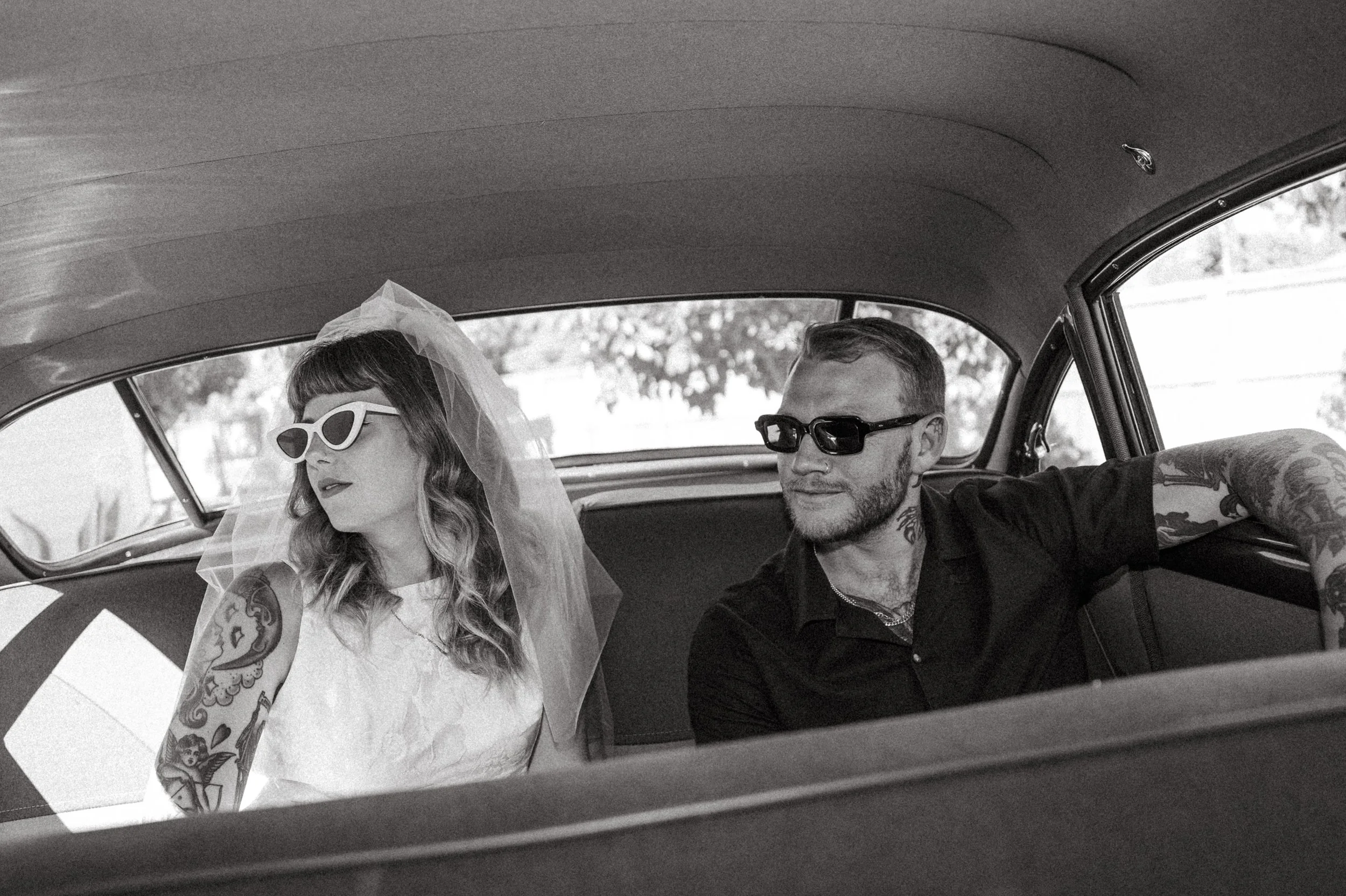 A man and woman sitting inside a vintage car. The woman is wearing a wedding dress, veil, and sunglasses, and has tattoos on her arm. The man is wearing sunglasses and a black shirt, and has tattoos on his arm and neck.