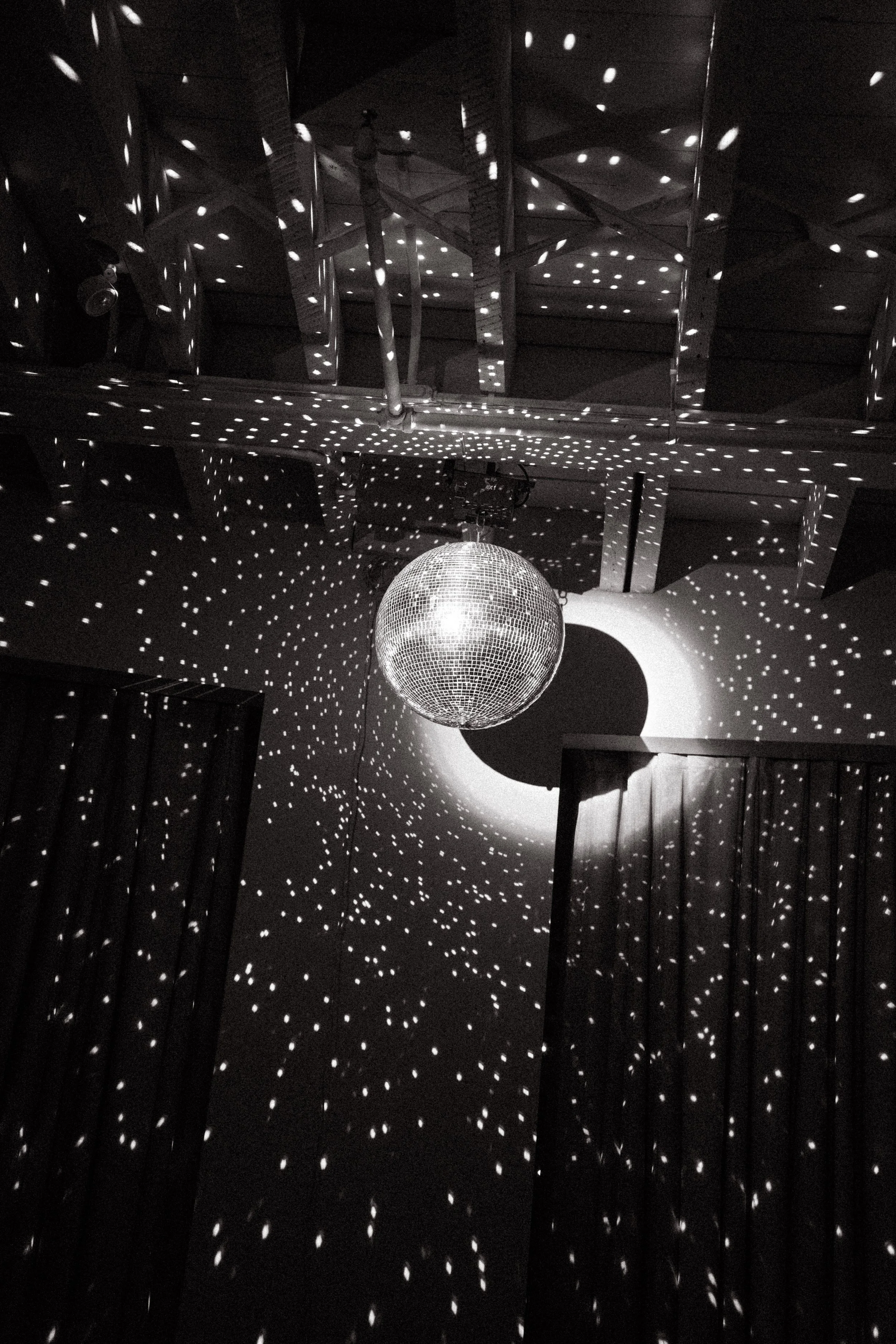 Black and white photo of a disco ball hanging from a ceiling, casting numerous small light reflections on the walls and curtains, creating a starry effect in the room.