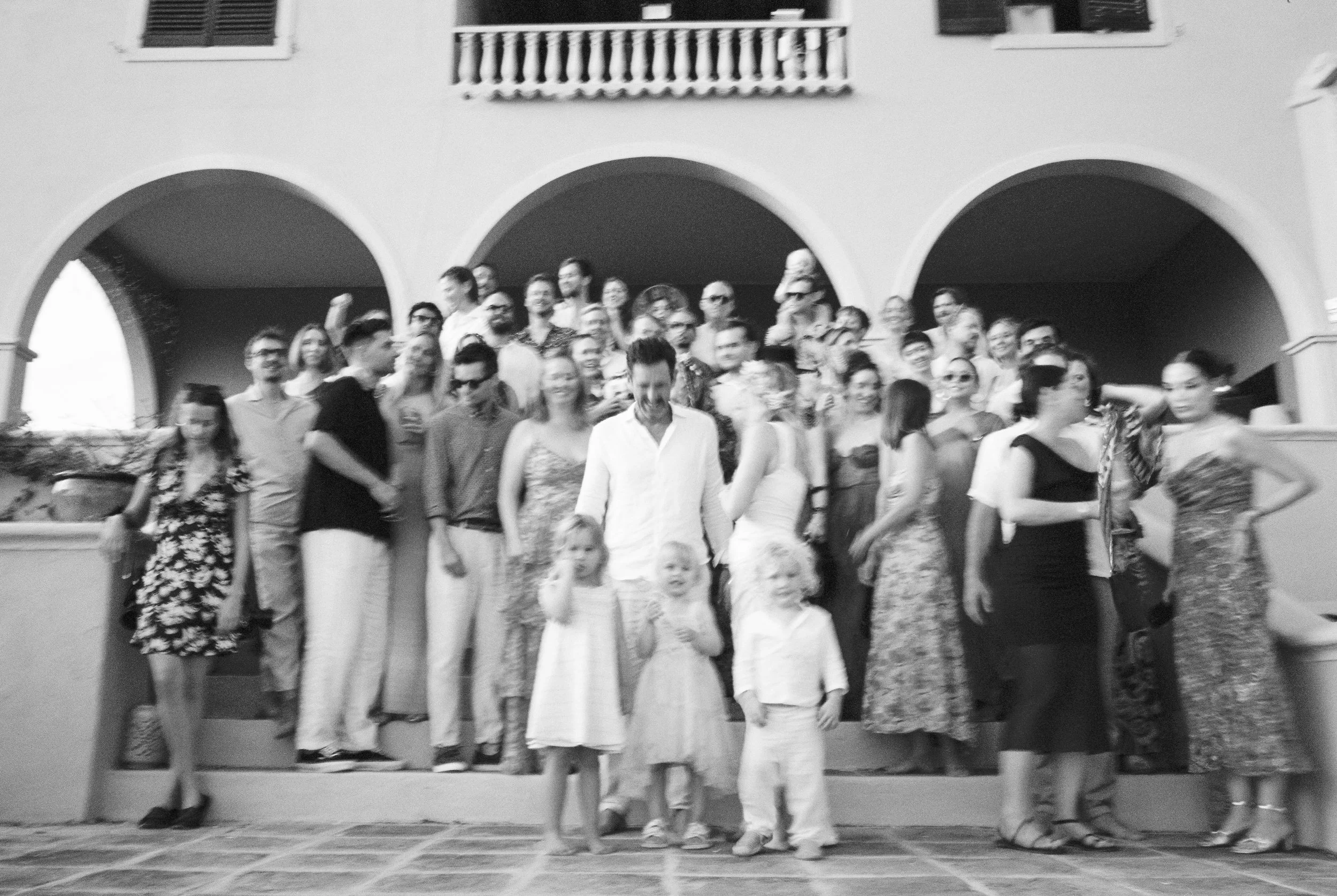 A large group of people, including children, gathered for a photo on the steps of a building with arches and balconies, in black and white.