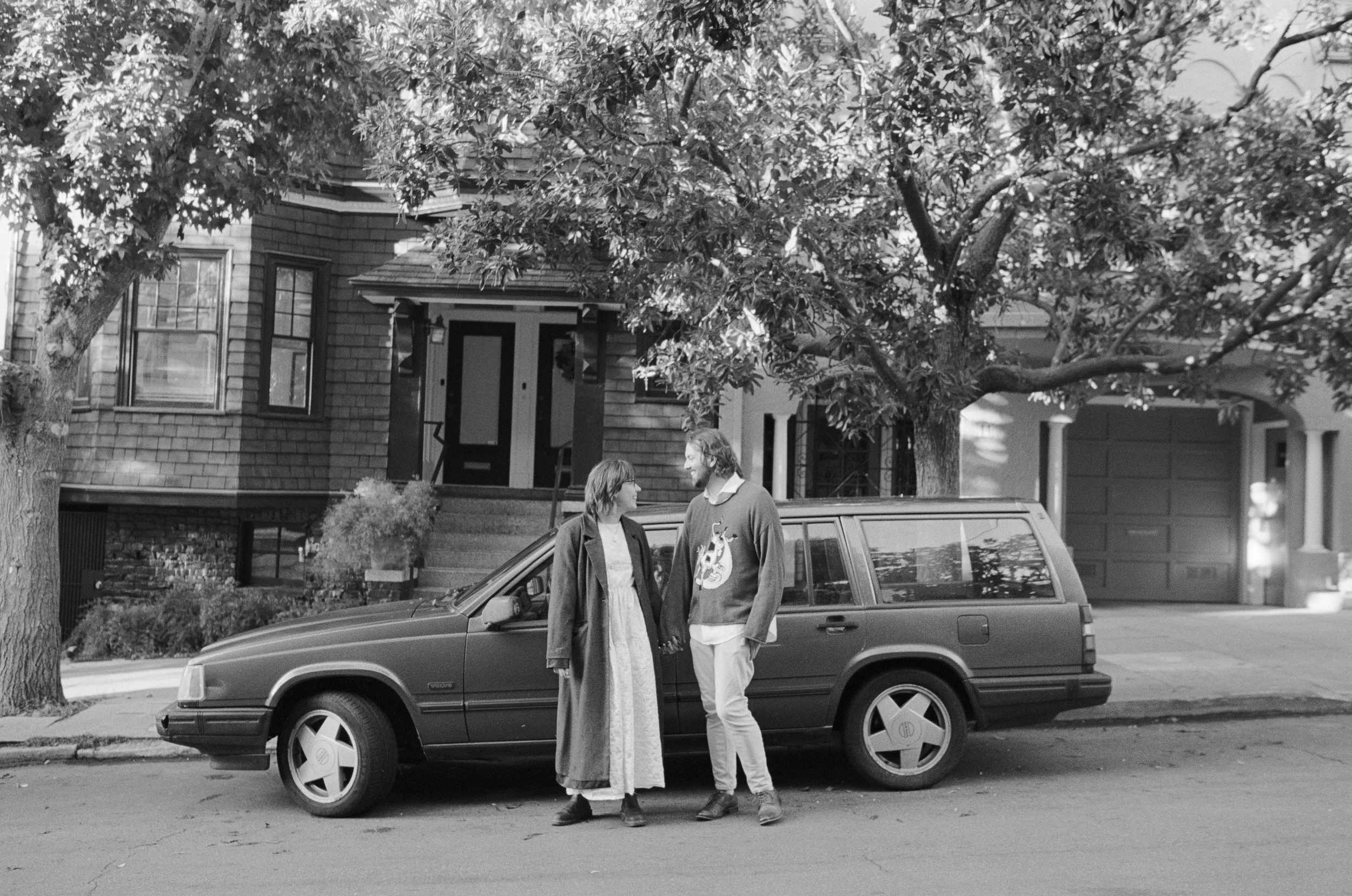 A black and white photo of two people standing and talking on a residential street next to a parked station wagon. There are trees and houses in the background.