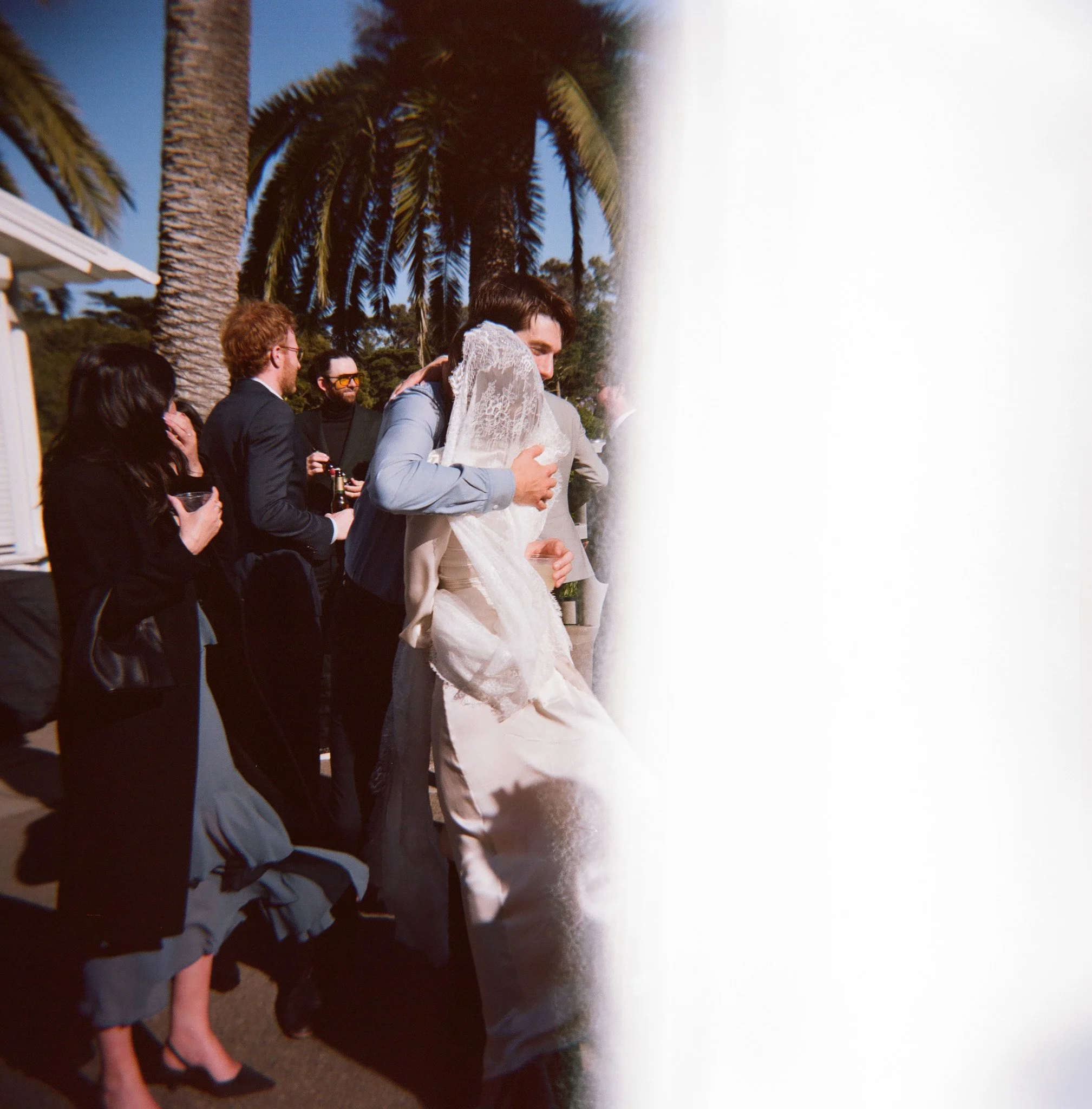 A wedding celebration outdoors with palm trees in the background. Two people are hugging, one in a suit and the other in a wedding dress. Several guests in formal attire are standing nearby, some holding drinks, under a clear blue sky.
