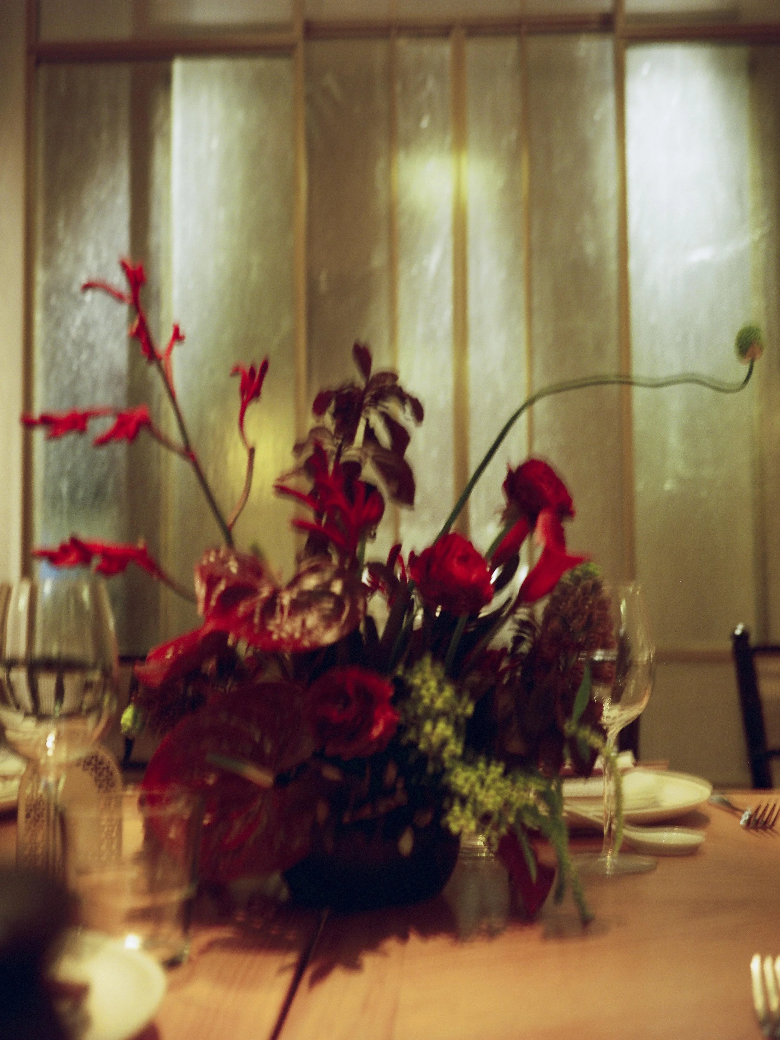 A floral centerpiece on a dining table with plates, forks, and glasses, set against a background of textured glass panels.